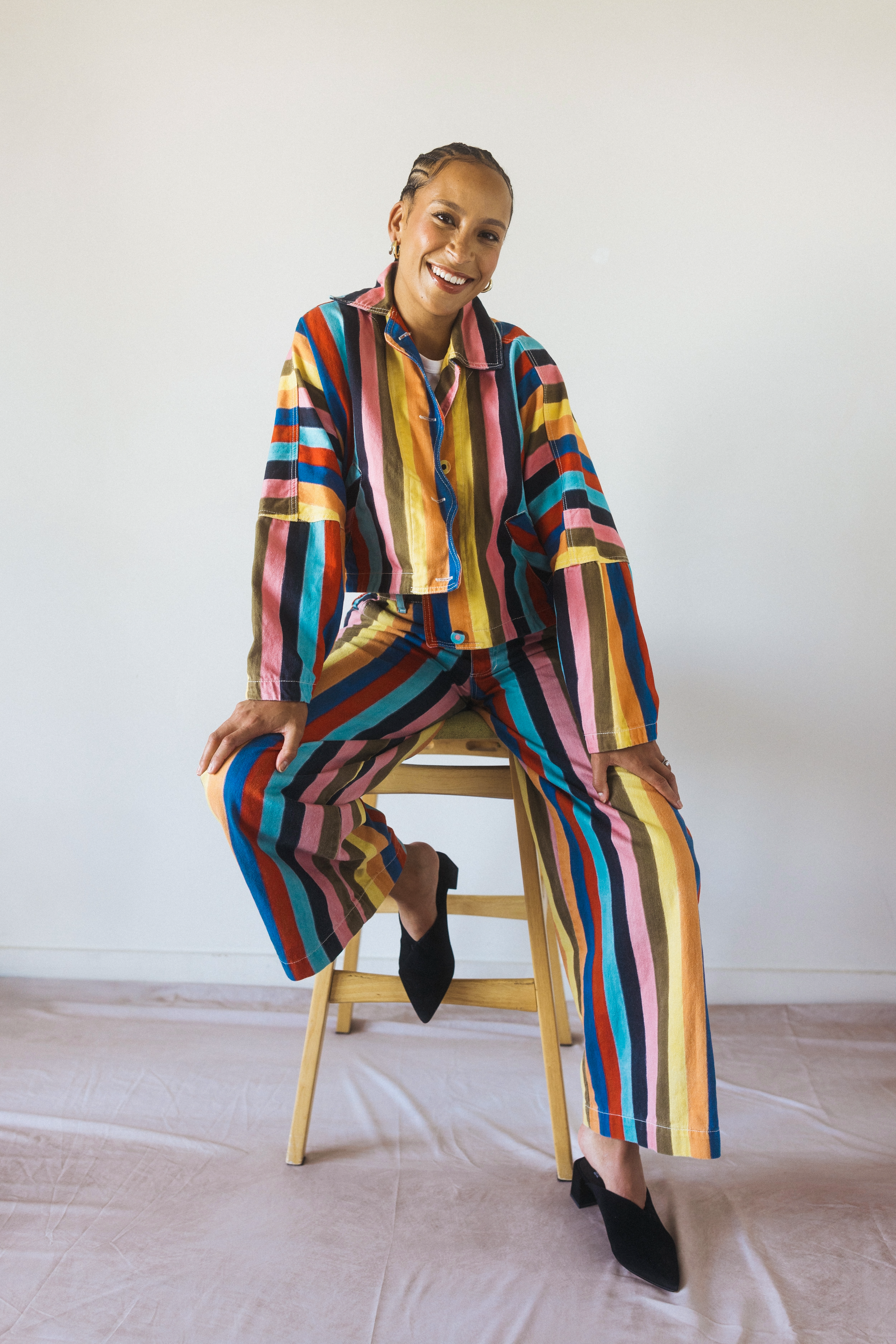 A woman wearing a colorful striped jacket and matching pants seated on a wooden chair with one foot resting on the chair's rung. She is smiling, has her hair styled in braids, and is wearing earrings, black high heels, and a white shirt under her jacket in a minimalist room with a plain white wall and a beige floor.