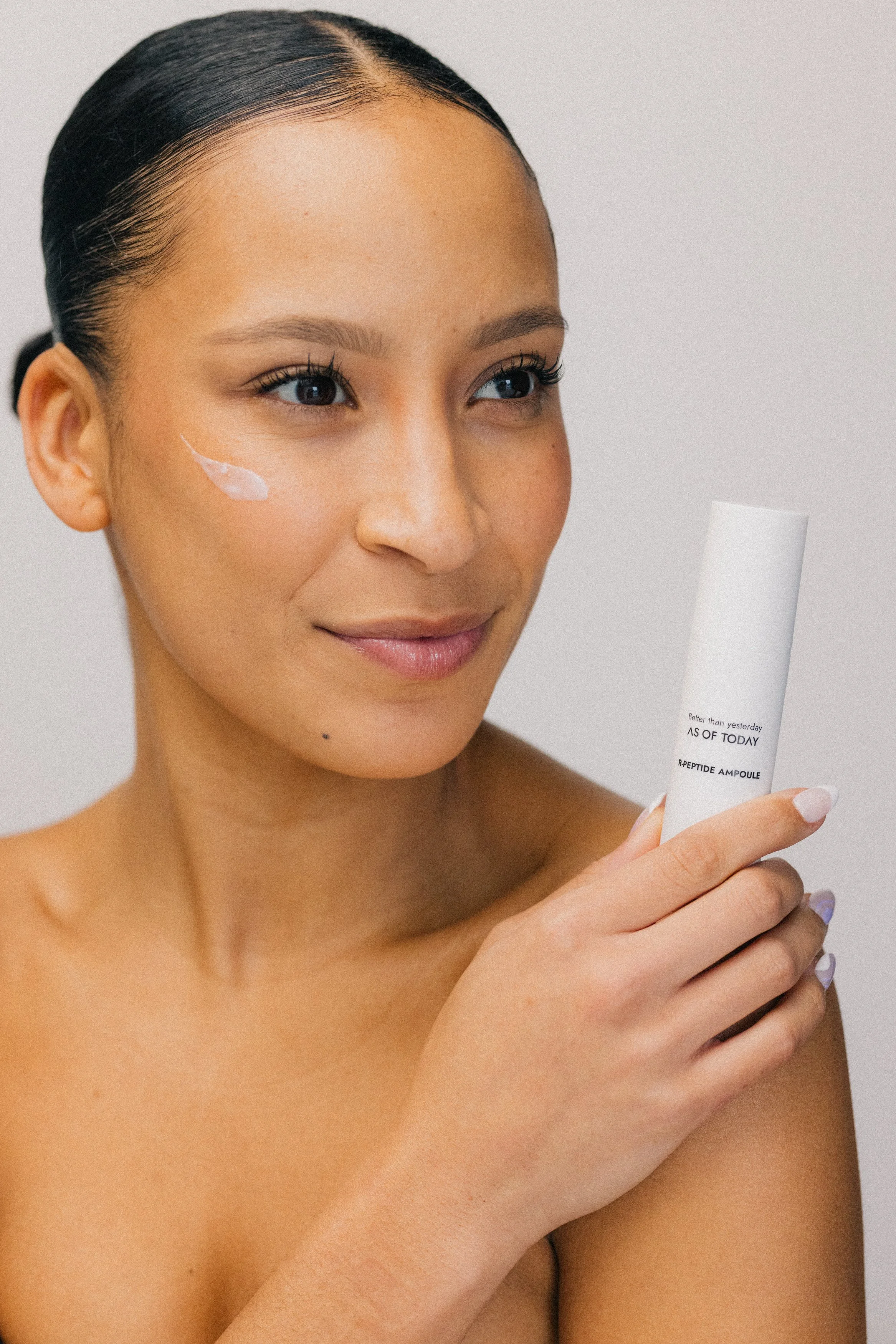 A woman with blue eyes and dark hair is holding a white skincare product with text on it. She has a small amount of cream on her face near her cheekbone, and she is smiling softly. The background is plain and light-colored.