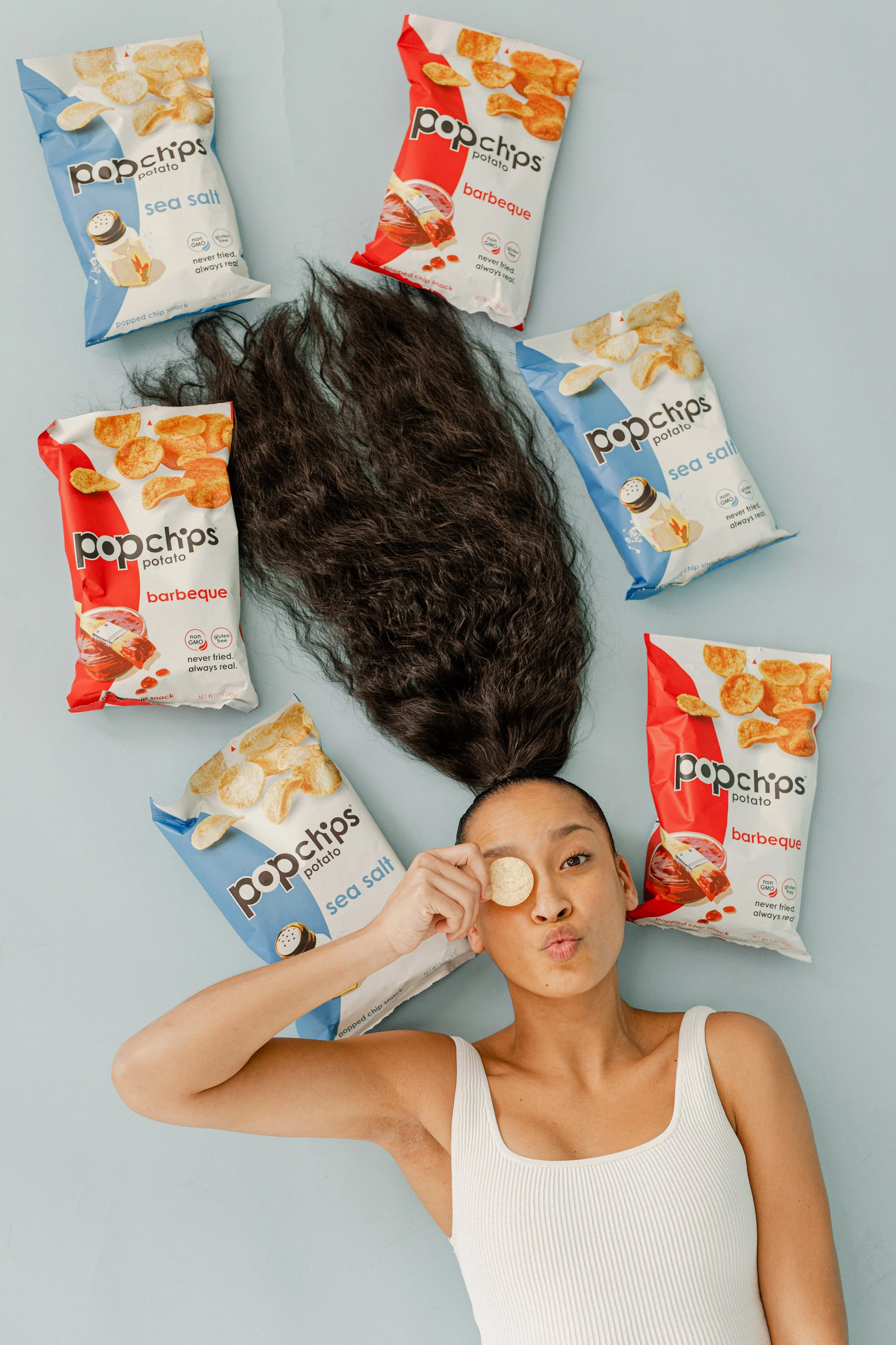 A woman lying on a light blue background with long brown hair, holding a potato chip up to her eye, surrounded by bags of Pop Chips potato chips in sea salt and barbeque flavors. She is wearing a white tank top and making a playful expression.