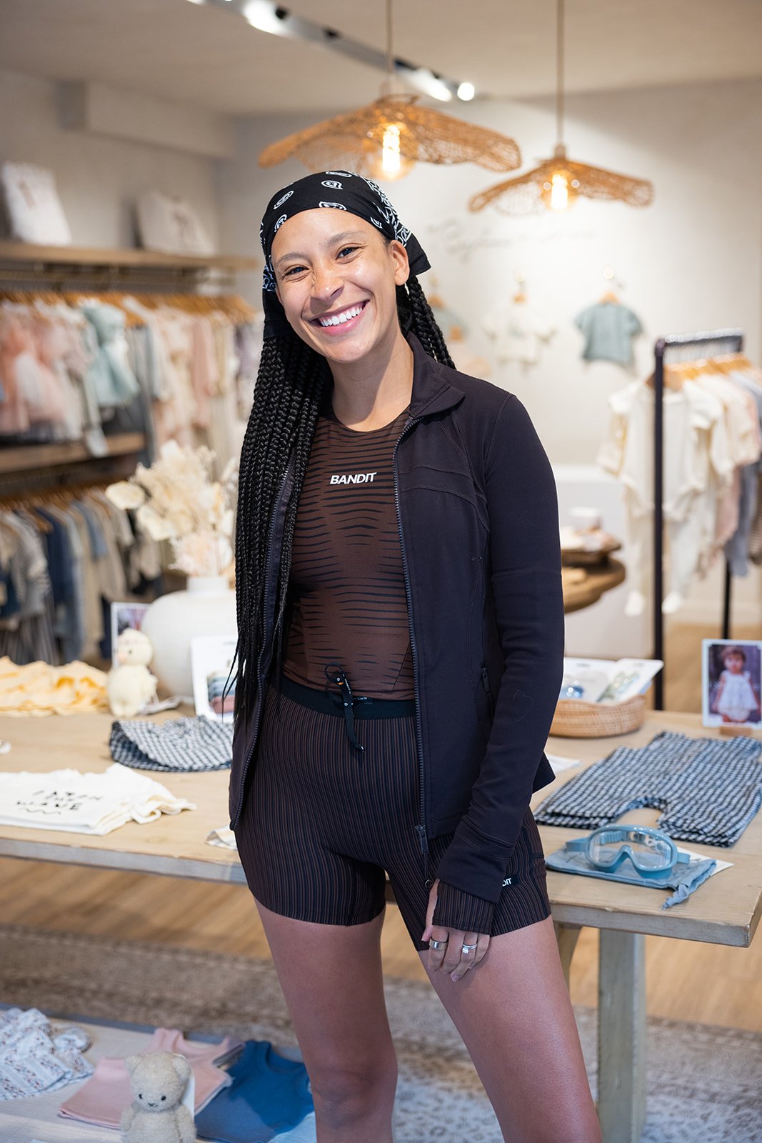 A woman with long braided hair wearing a black bandana, black jacket, and striped black shorts, smiling in a boutique clothing store with racks of clothes and accessories in the background.
