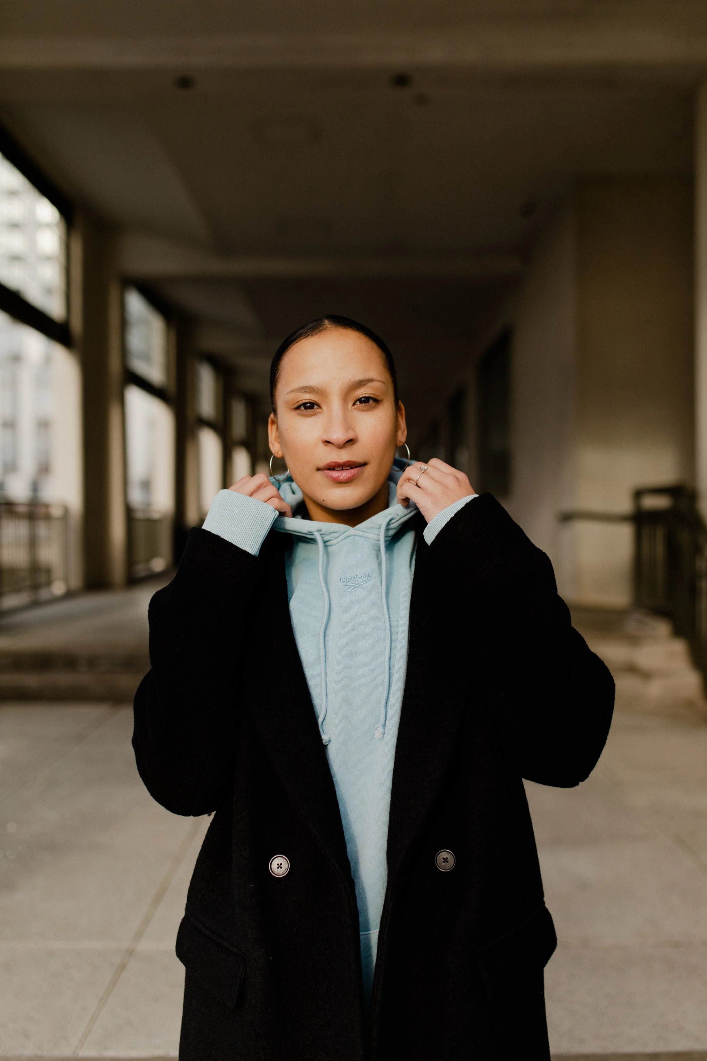 Young woman with dark hair pulled back, wearing large hoop earrings, a light blue hoodie, and a black coat, standing in an urban corridor with windows and concrete walls.