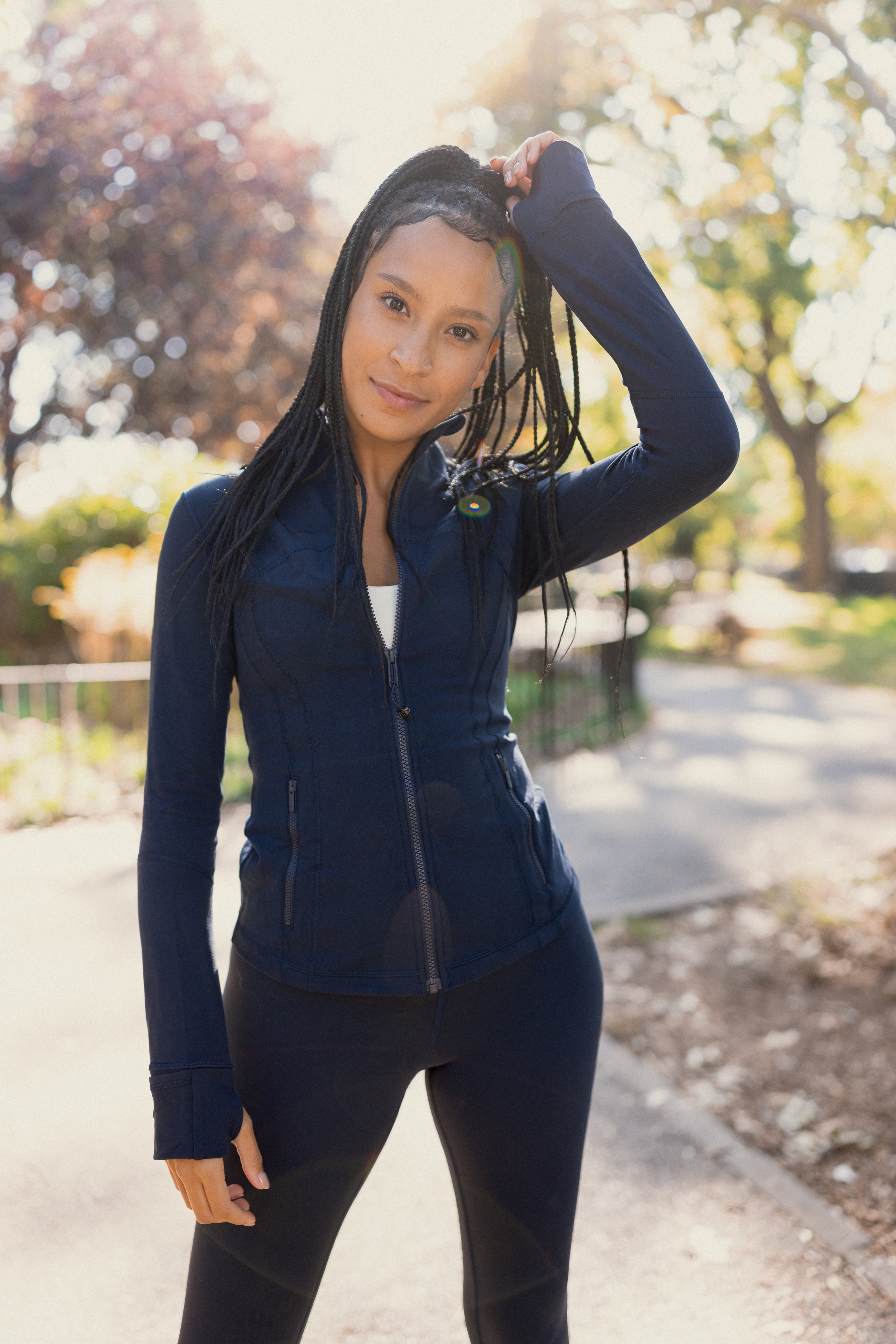 A woman in black athletic clothing standing outdoors on a tree-lined path during autumn, with trees showing fall foliage in the background.