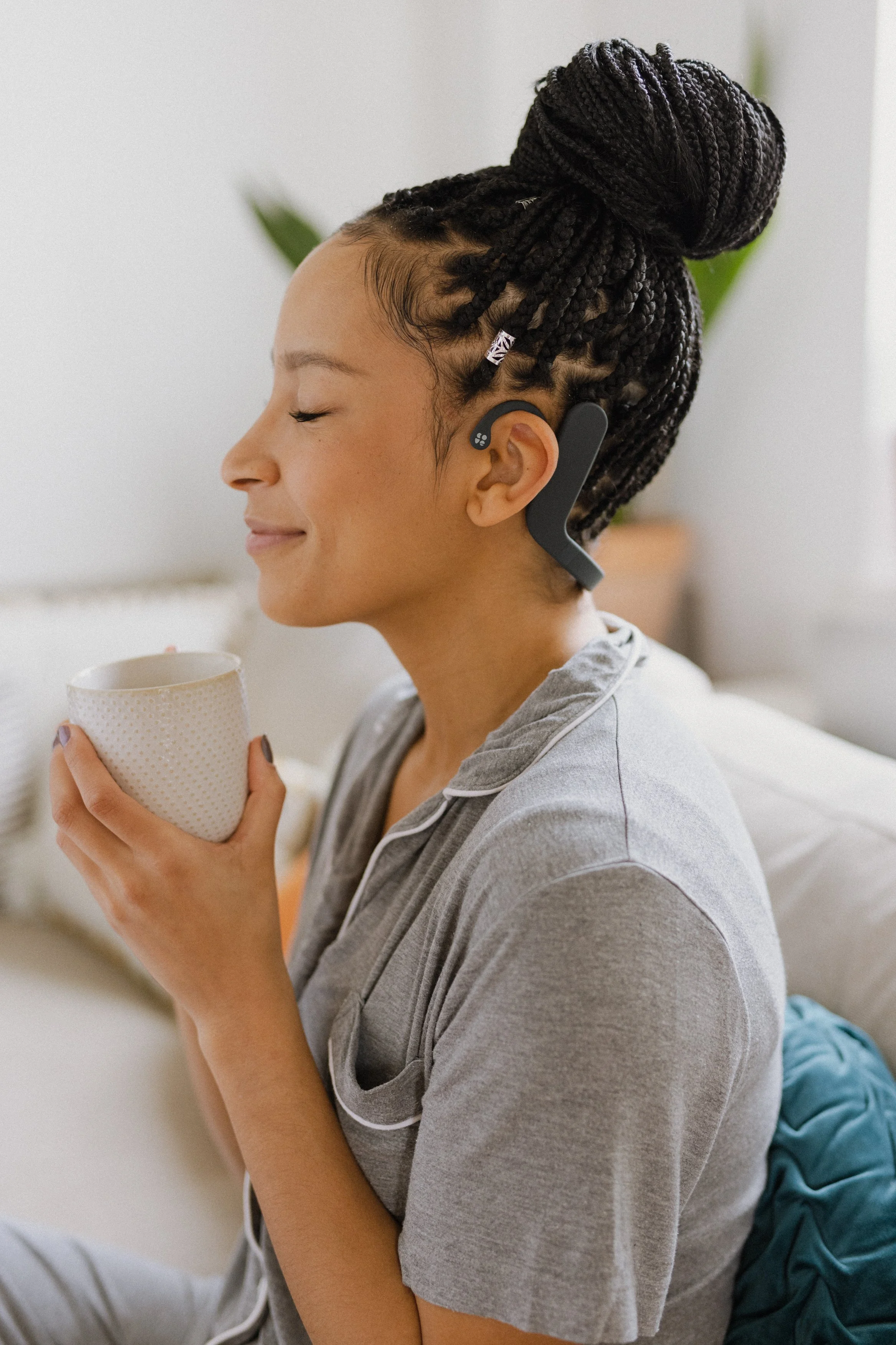 A young woman with braids in her hair, smiling with closed eyes, holding a white mug, sitting on a bed with a light-colored wall and a potted plant in the background.