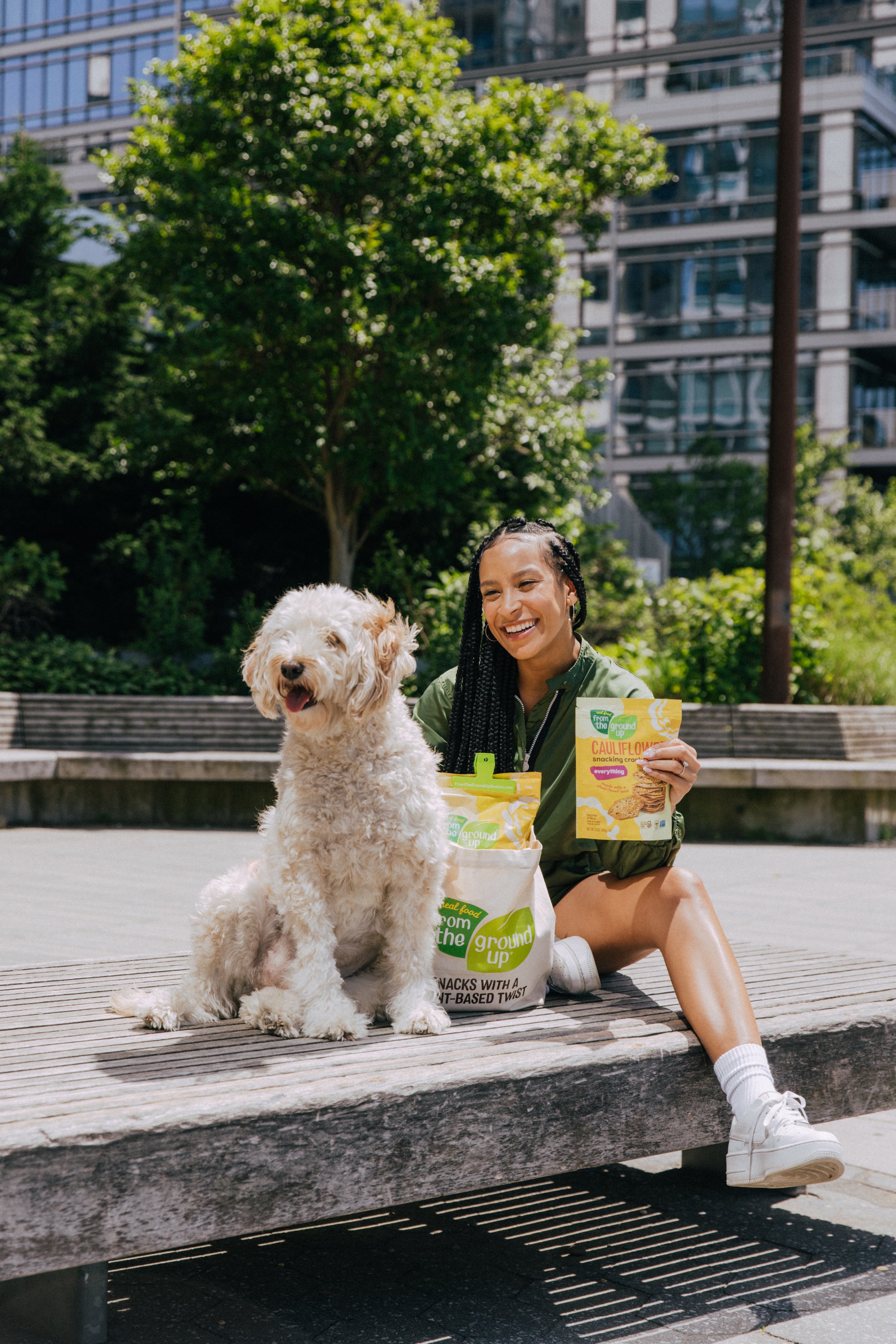 A young woman with braided hair sitting on a park bench, smiling, with a large fluffy white dog next to her. She is holding a bag of Cauliflower snack crackers. The background features trees and modern buildings.