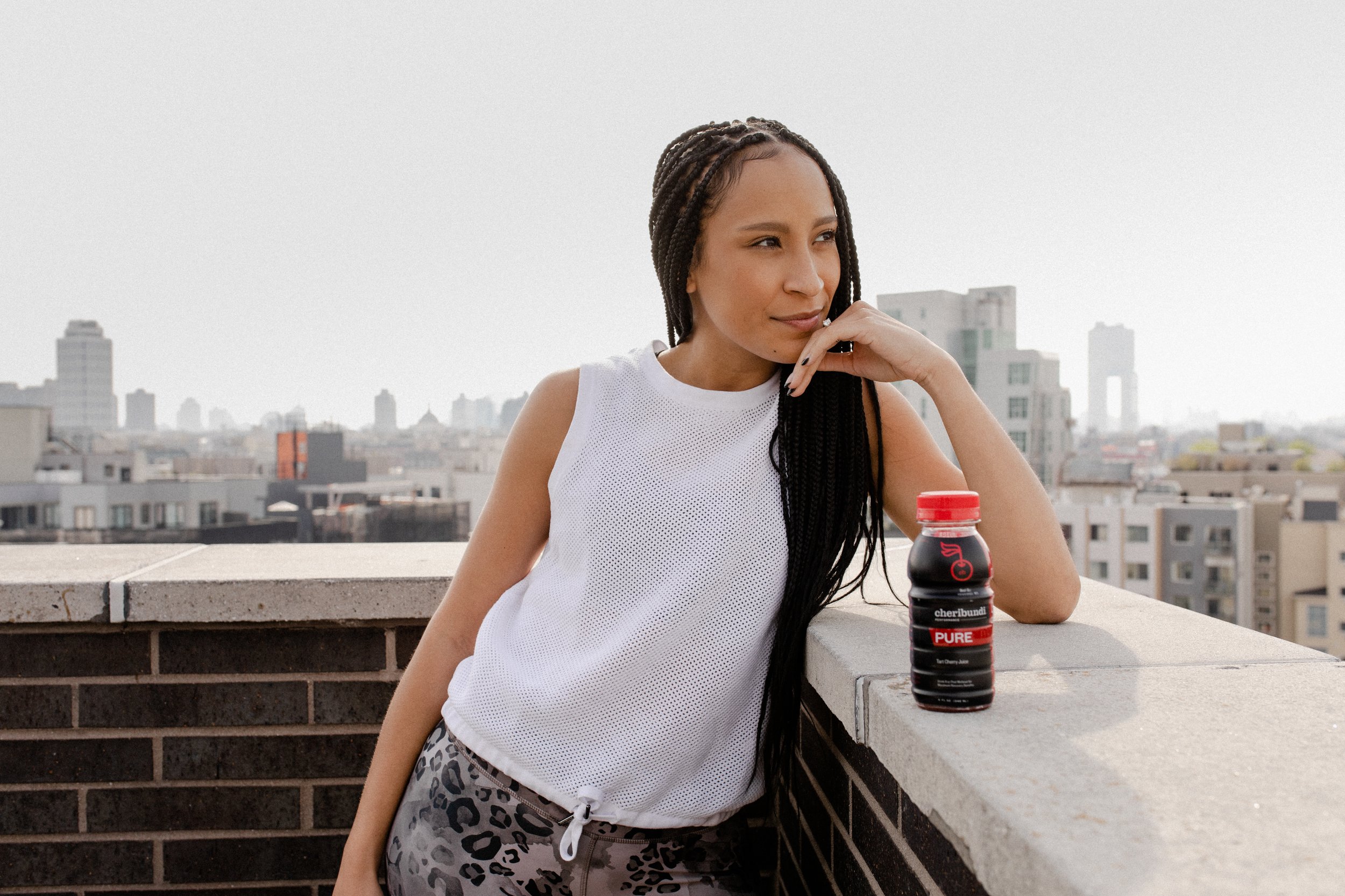 A woman with long braided hair is leaning on a rooftop ledge with her hand near her face. She is wearing a white sleeveless top and leopard print shorts. There is a bottle of Cheribundi juice on the ledge in front of her. The city skyline is in the b