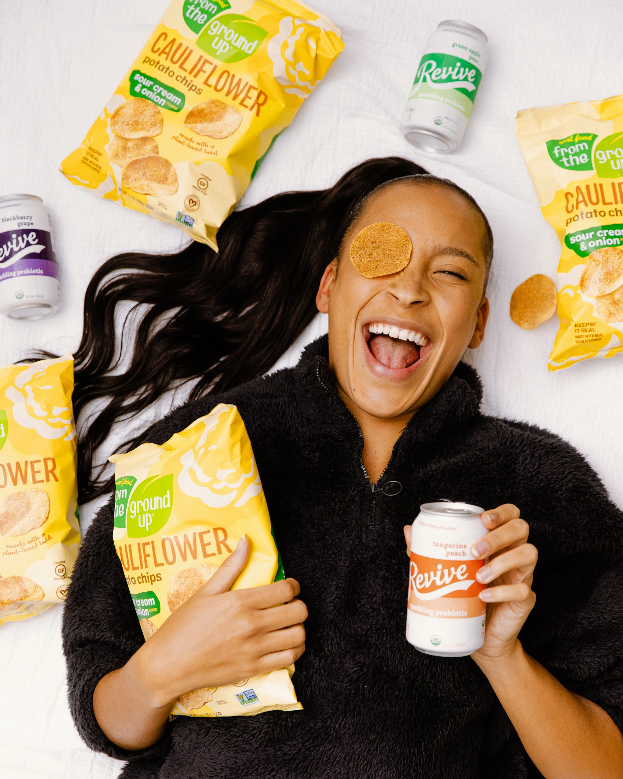 A smiling woman with long dark hair lying on a white surface, surrounded by yellow bags of cauliflower potato chips, holding a white can of Revive probiotic beverage with peach and tangerine flavor, with one chip placed on her forehead.