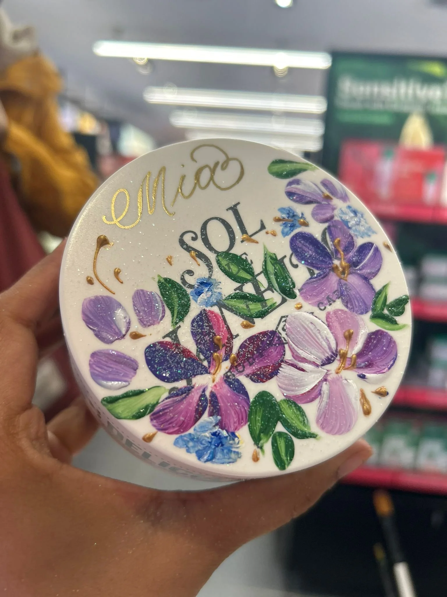 A hand holding a container of Sol de Janeiro body cream with floral design, purple and pink flowers, green leaves, and gold accents, with the brand name and product description partially visible.