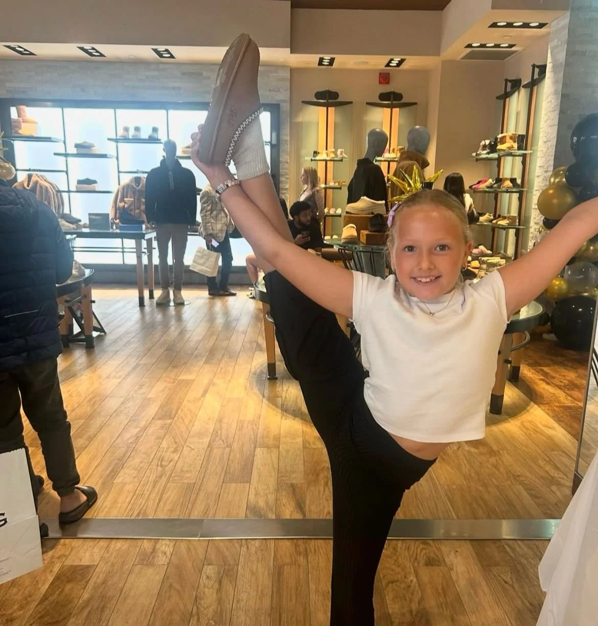 A young girl in a white t-shirt and black pants is smiling and holding one leg up high, balancing on one foot inside a shoe store. The store has wooden floors, shelves with shoes, and other shoppers in the background.