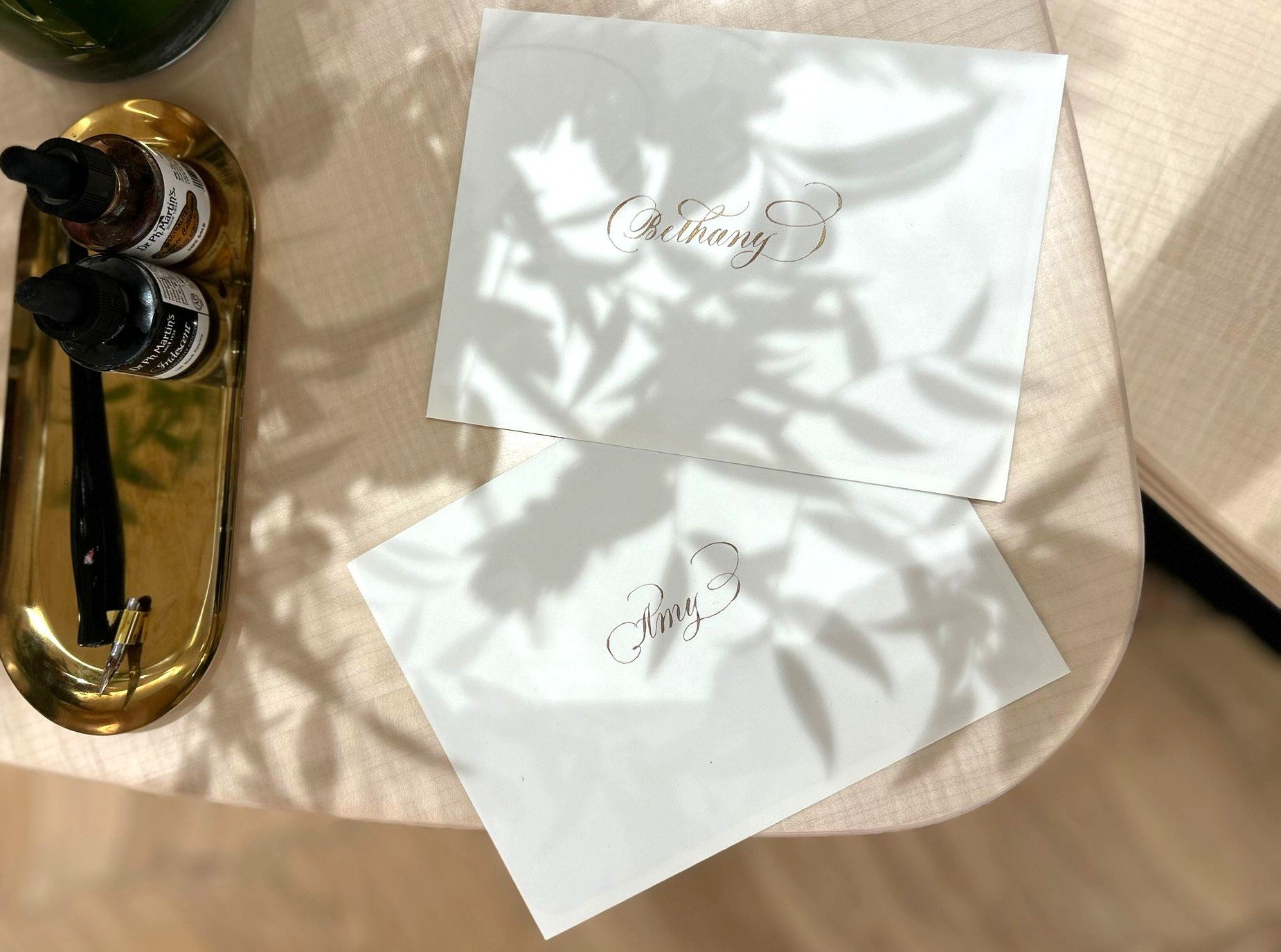 Two white pieces of paper on a beige table, with elegant cursive writing that reads "Bethany" and "Amy 3". Shadows of plant leaves cast across the papers. To the left, a gold tray with small bottles and a pen.