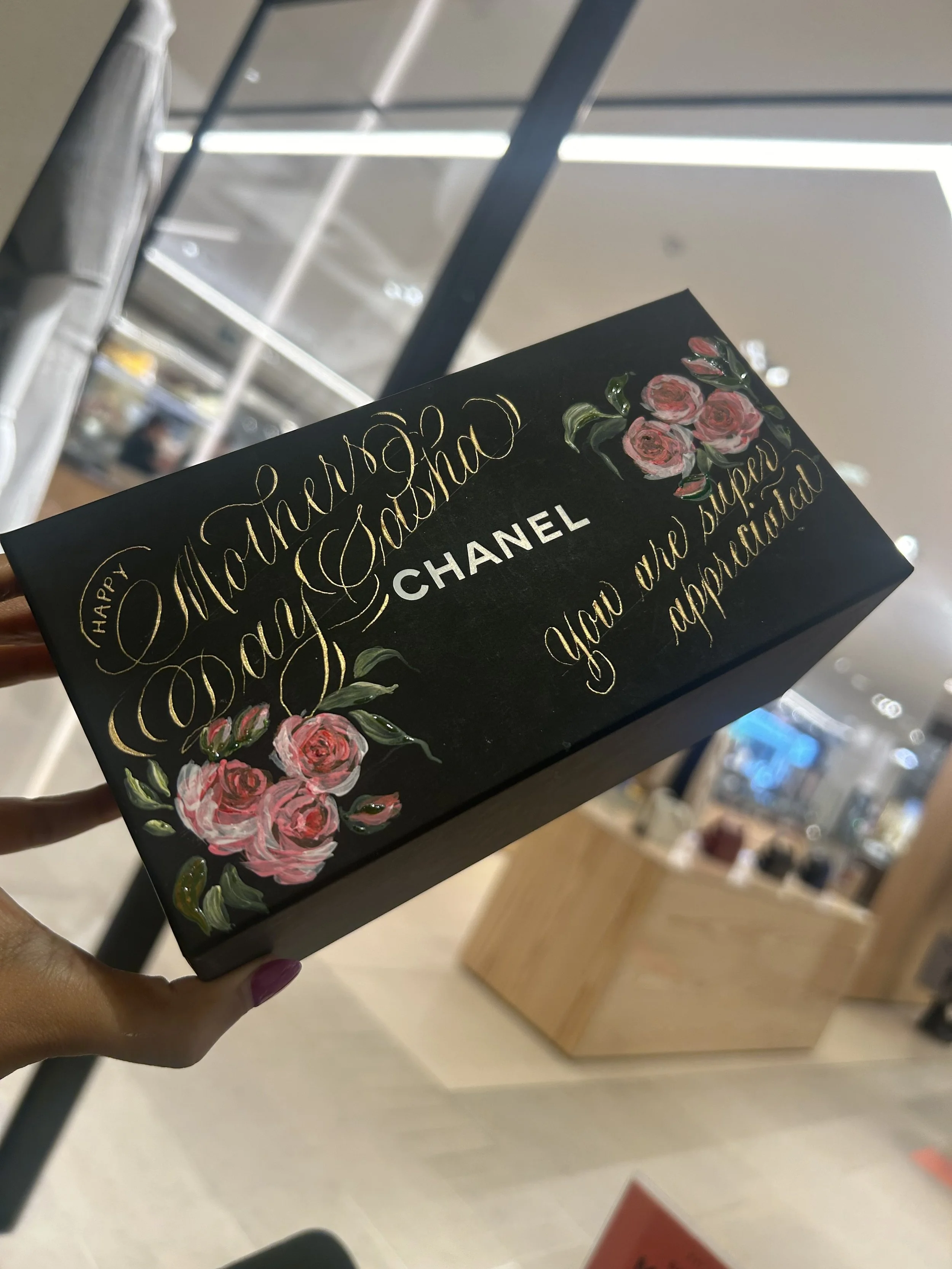 Black gift box with gold and white lettering and pink floral decorations, reading 'Happy Mother’s Day, you are loved and appreciated' and featuring the Chanel logo.