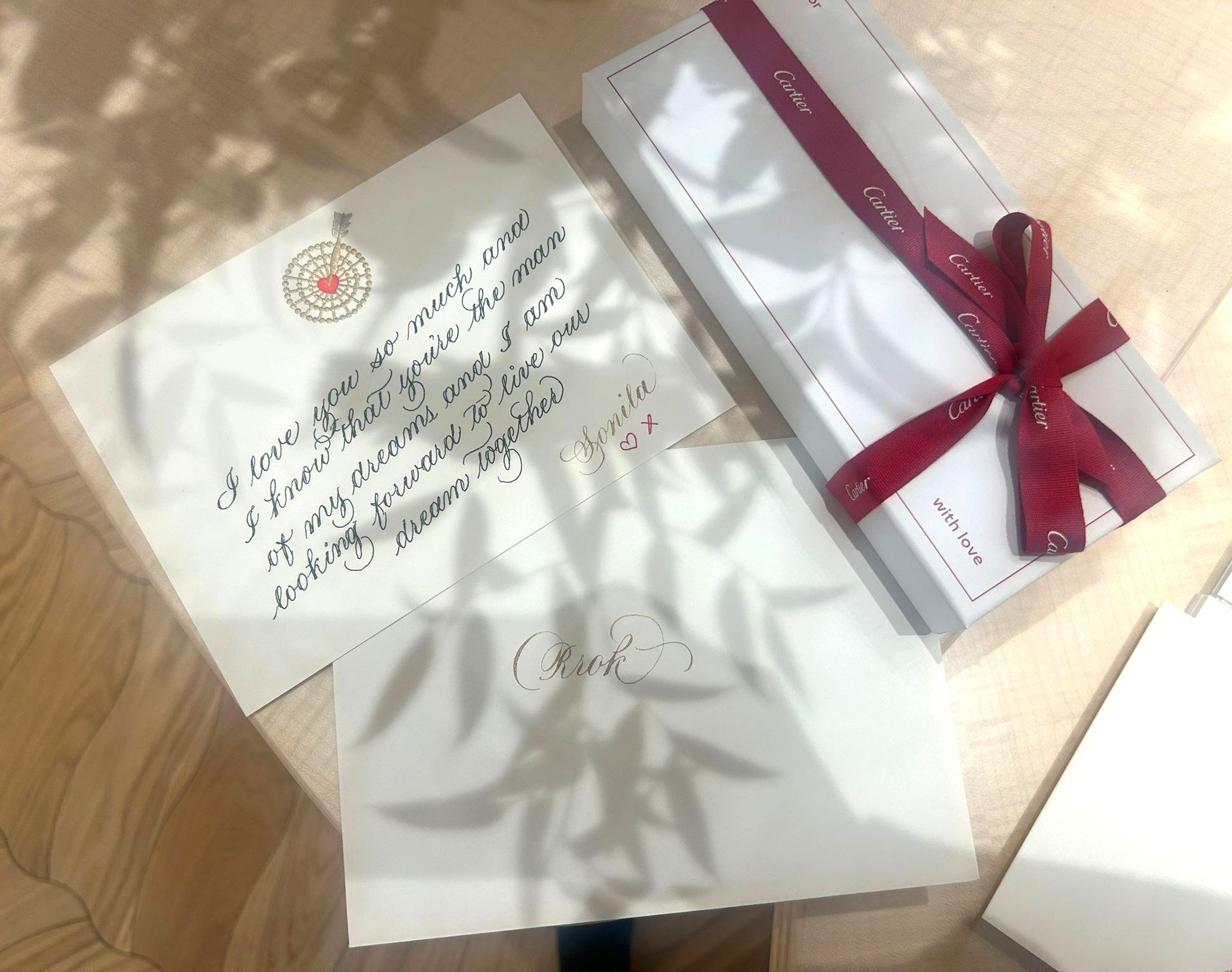A handwritten note and a box of Cartier chocolates on a table, surrounded by shadows of leaves.