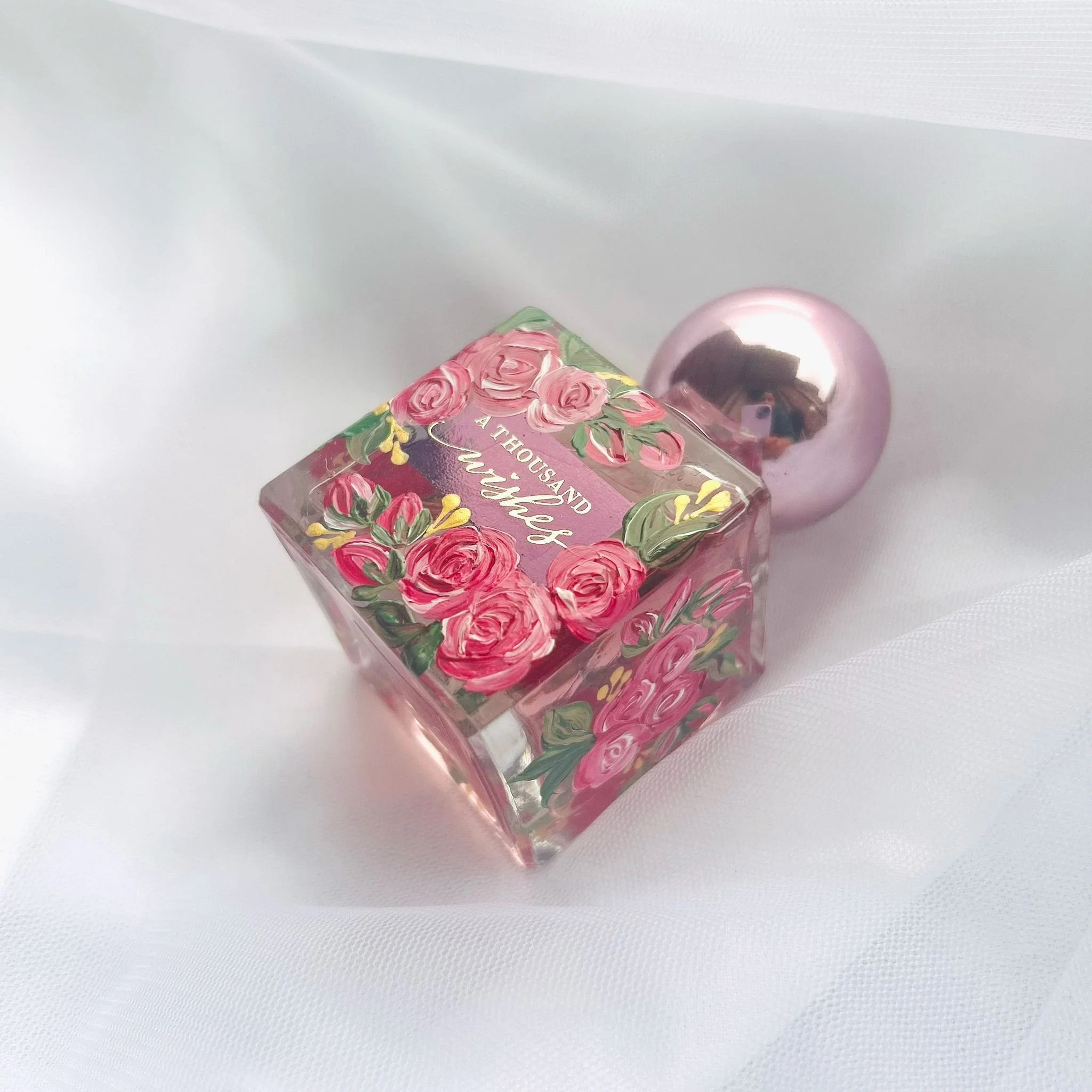 A glass perfume bottle with pink roses and the words 'A THOUSAND WISHES' on the front, resting on a white fabric surface.