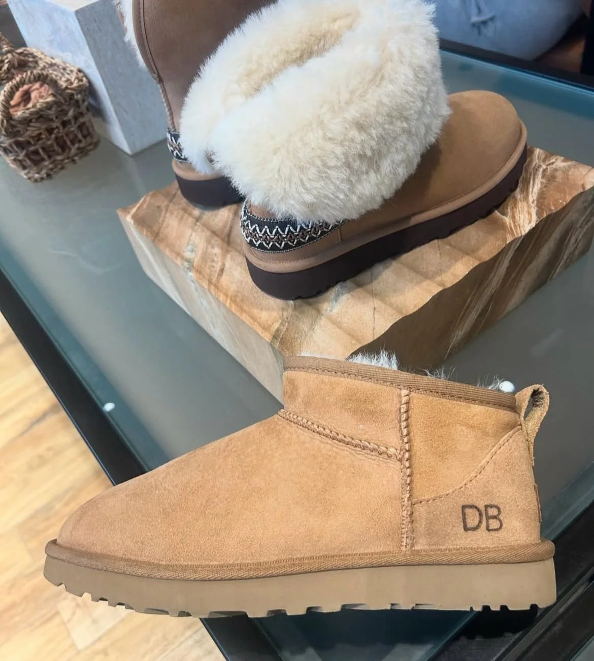 Close-up of a tan UGG-style sheepskin boot with the initials 'DB' embroidered on the side, placed on a glass display table.