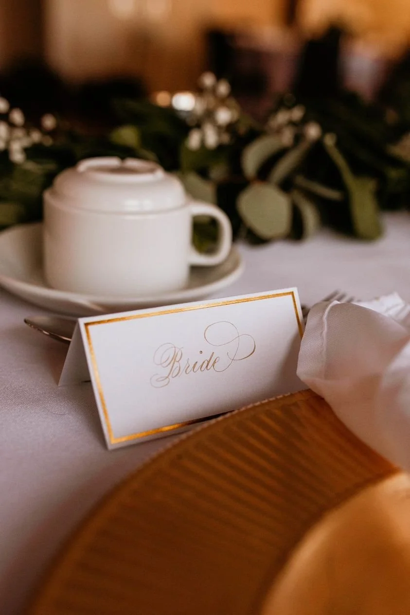 A table setting at a wedding reception with a white place card labeled 'Bride' in gold cursive, a napkin, a white teacup on a saucer, and floral decorations in the background.