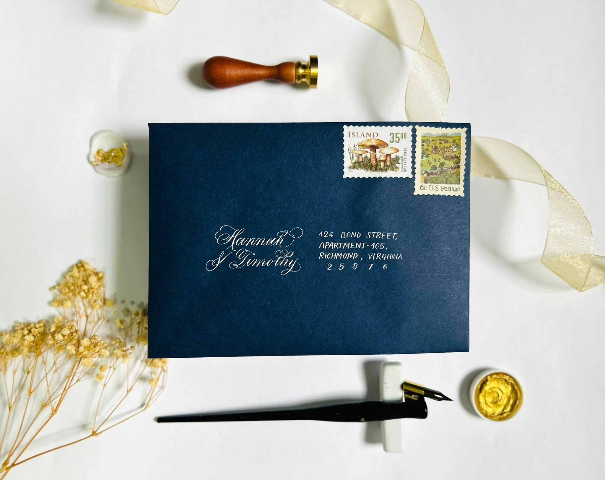 A navy blue envelope with elegant white calligraphy addressing Hannah and Timothy, decorated with vintage stamps, a sealing wax stamp, a dipping pen, and dried flowers, on a white background with a gold ribbon.
