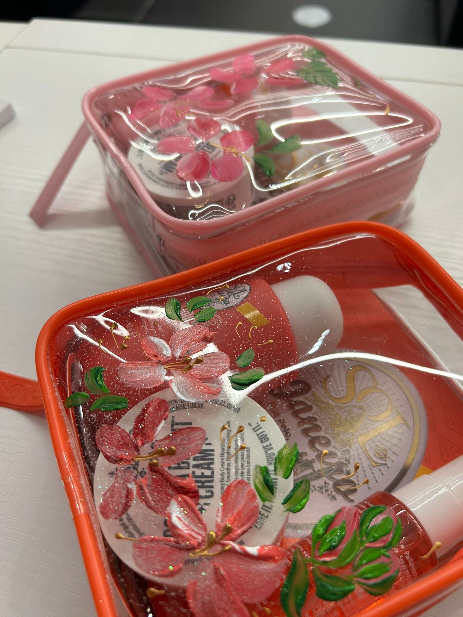 Close-up of two pink cosmetic containers with clear lids, decorated with flower and leaf patterns, placed on a white surface.