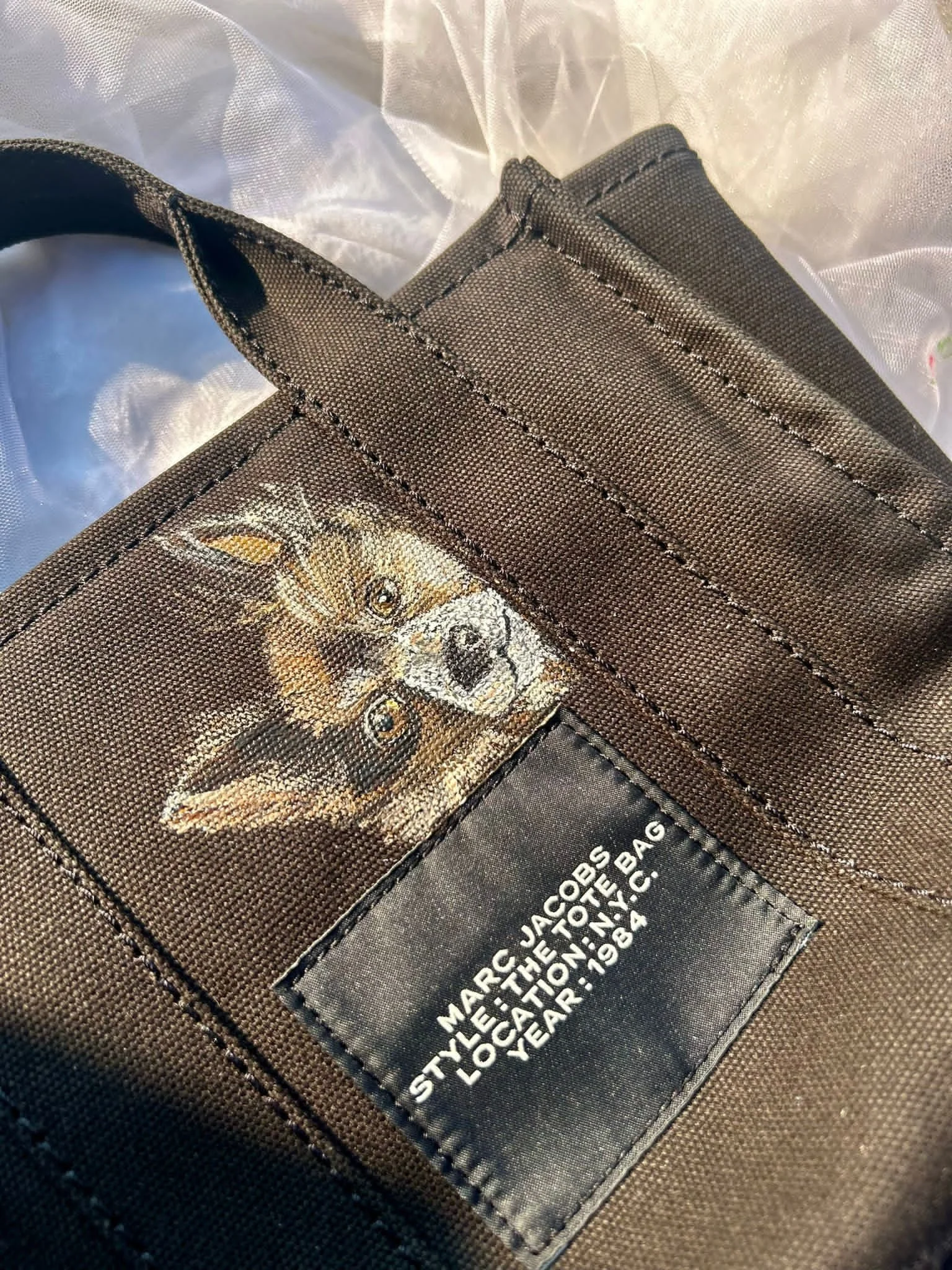 Close-up of a brown textured fabric bag showing a printed image of a dog and a black label with white text that reads 'MARC JACOBS THE TOTE BAG, L.O.CATION INC., YR: 1984'.