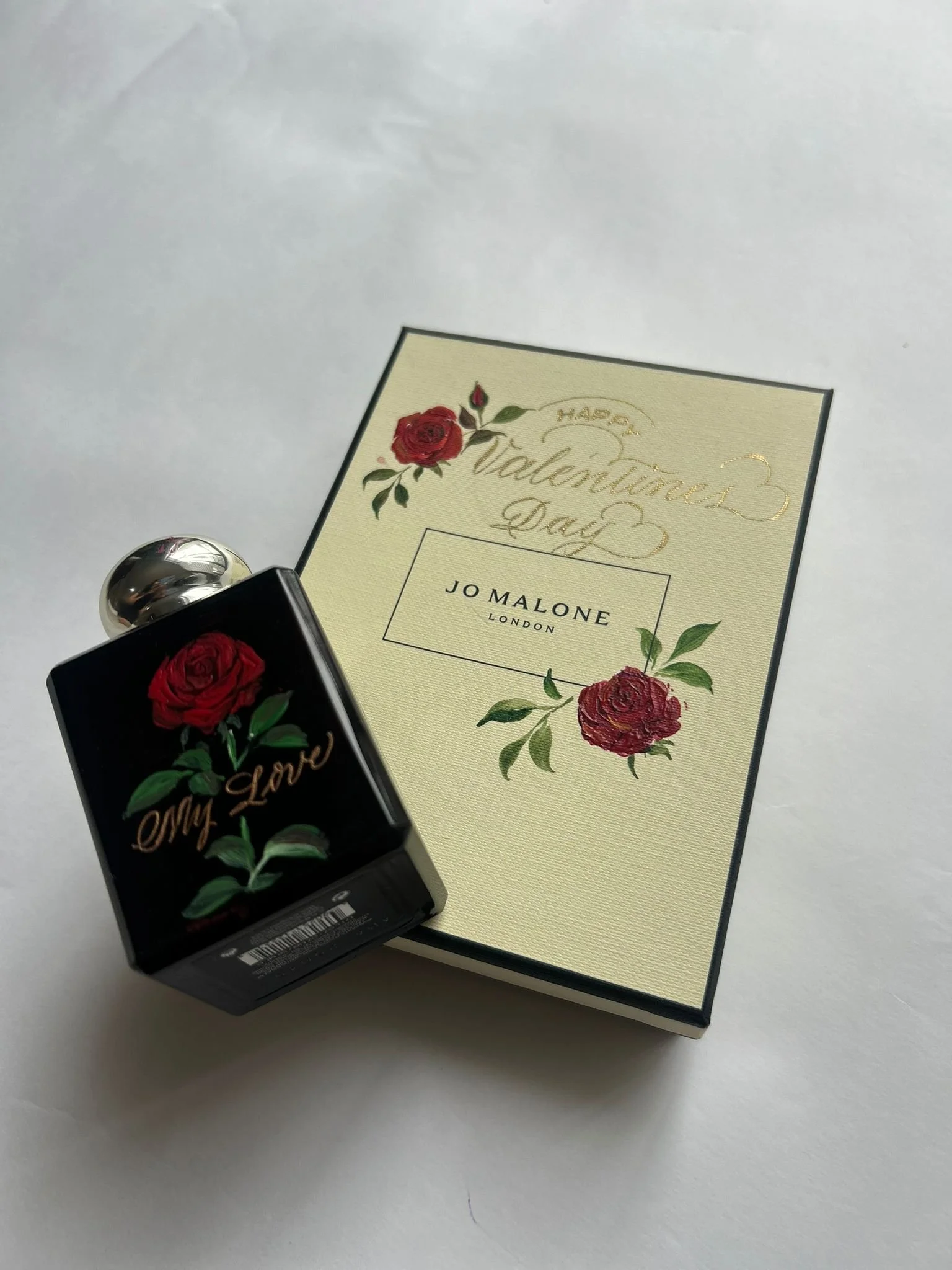 A perfume bottle with a black background and floral design, positioned next to a card with romantic roses and gold lettering, celebrating Valentine's Day.