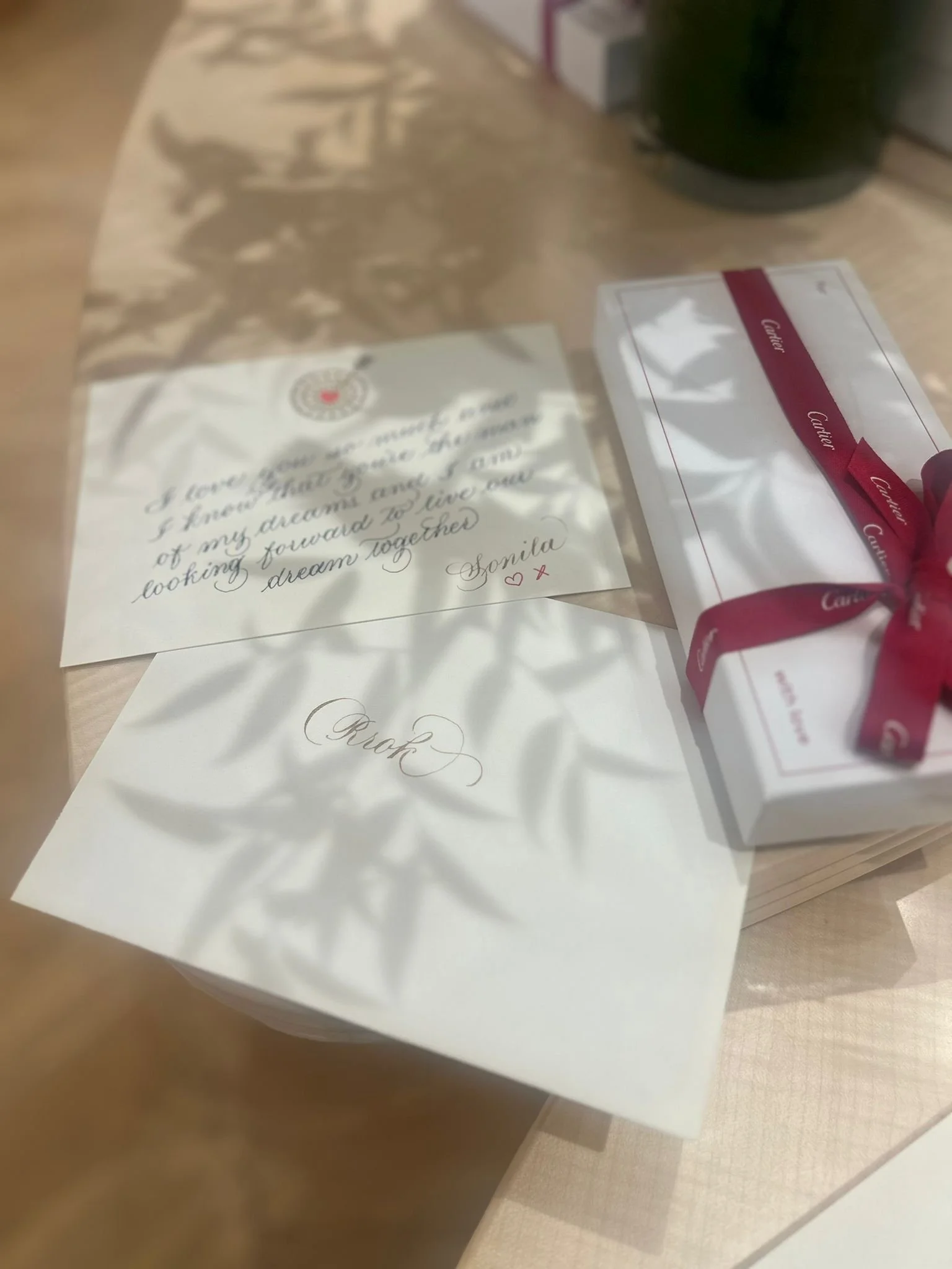 A handwritten note and a white box with a pink ribbon on a wooden surface, with a shadow of leaves cast on the note.