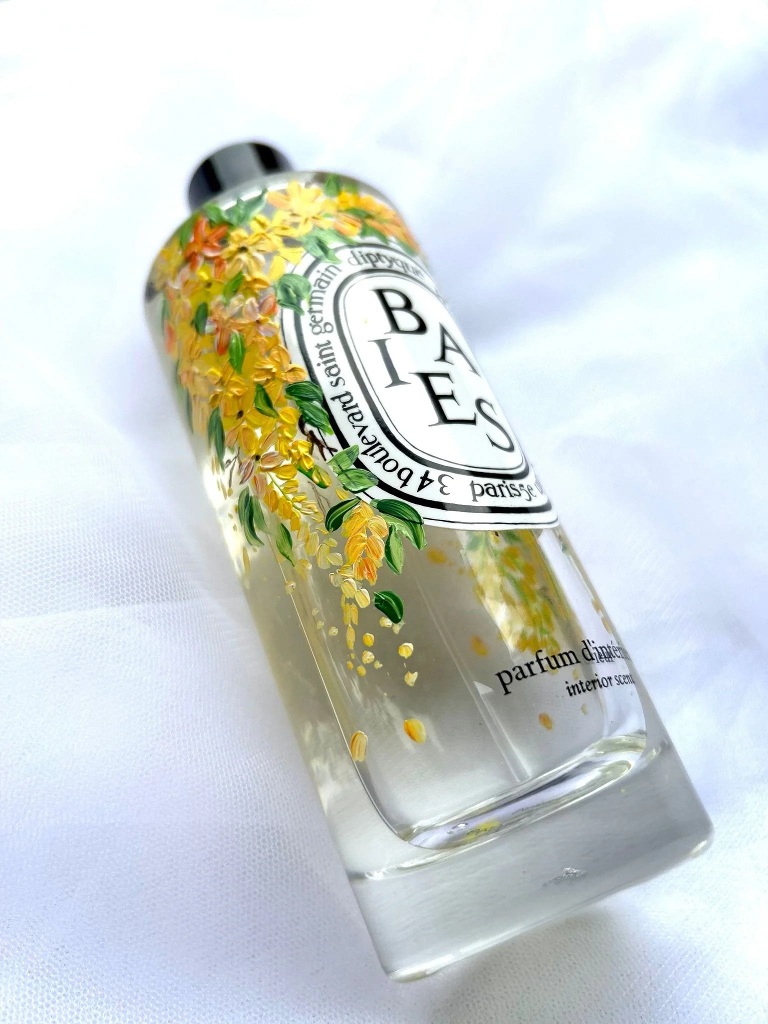 Glass perfume bottle with a white label, decorated with yellow flowers and green leaves, lying on a white surface.