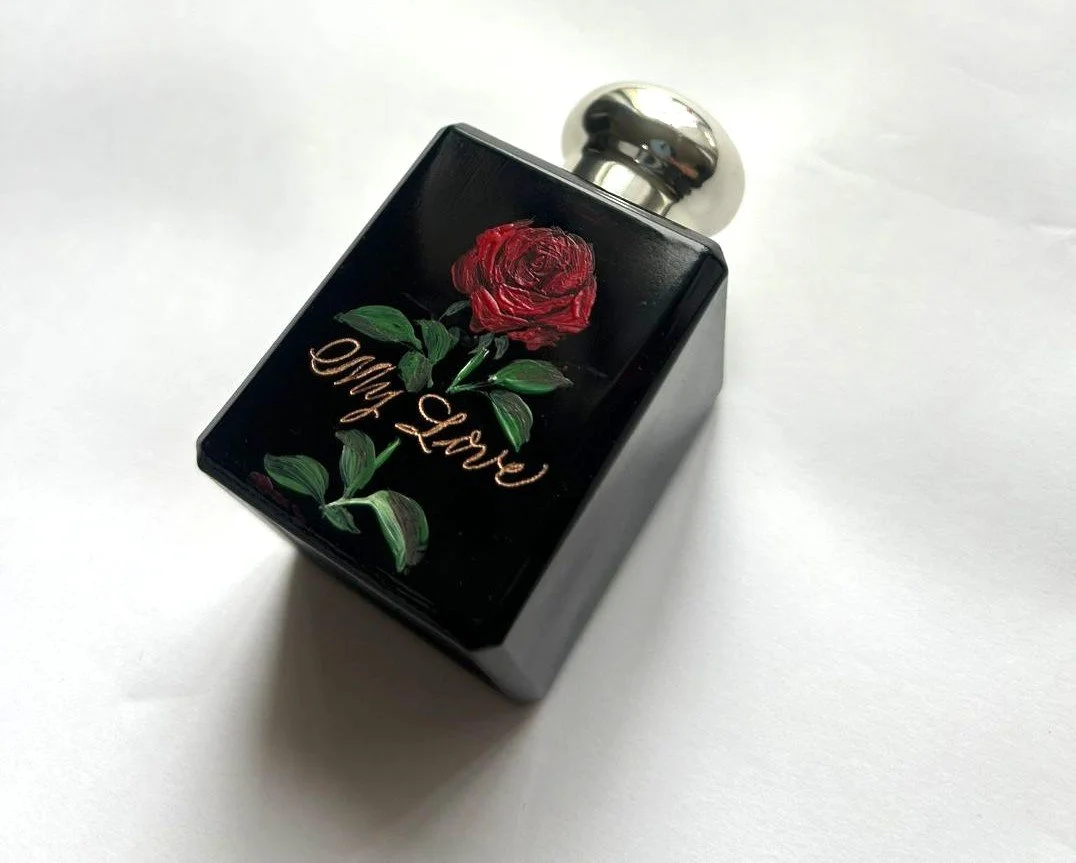 A black perfume bottle with a silver cap, decorated with a red rose and green leaves, and the words 'Amour' and 'Paris' written on it.