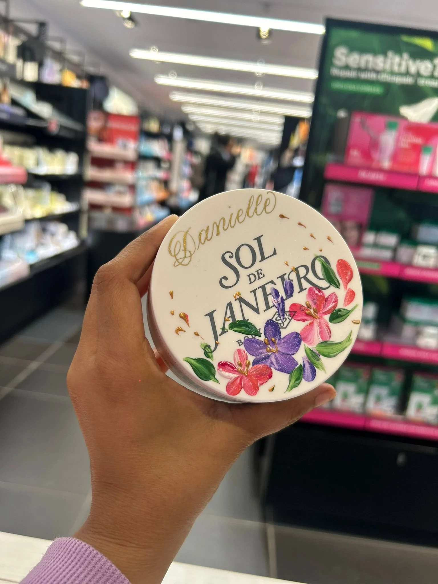A hand holding a round container with floral decoration and the message "Danielle Sol de Janiero" in a store aisle.
