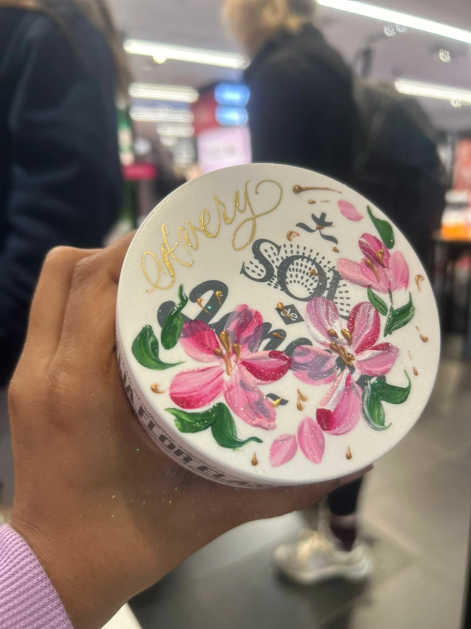 A person's hand holding a white container with floral artwork, pink flowers, green leaves, and gold accents, and the words "happy" and "social" visible on the lid. The background shows a blurred indoor setting with people and lights.
