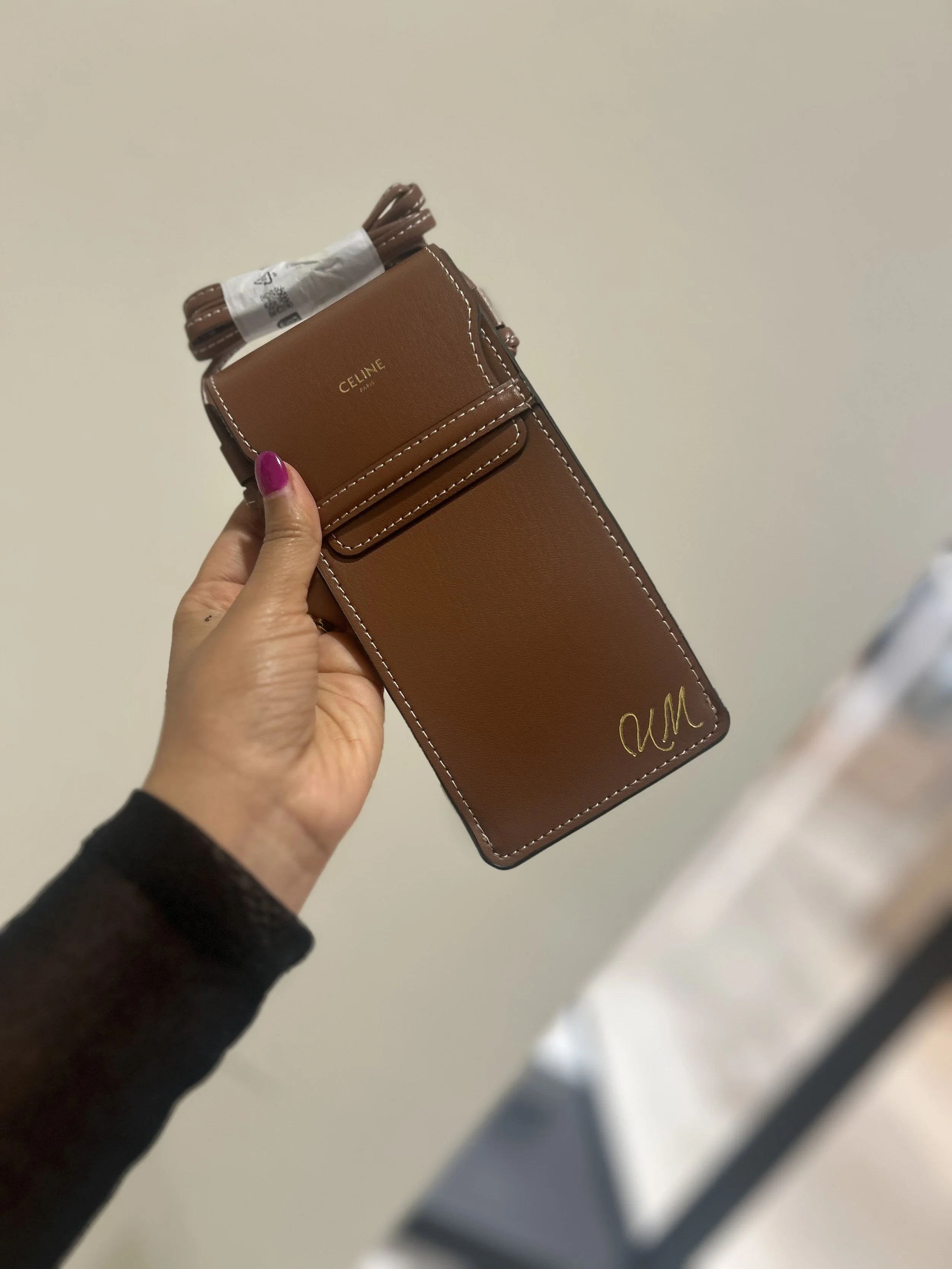 A person holding a brown Celine leather wallet with white stitching, featuring a signature on the bottom right corner and a small flap pocket on the front, against a plain beige background.