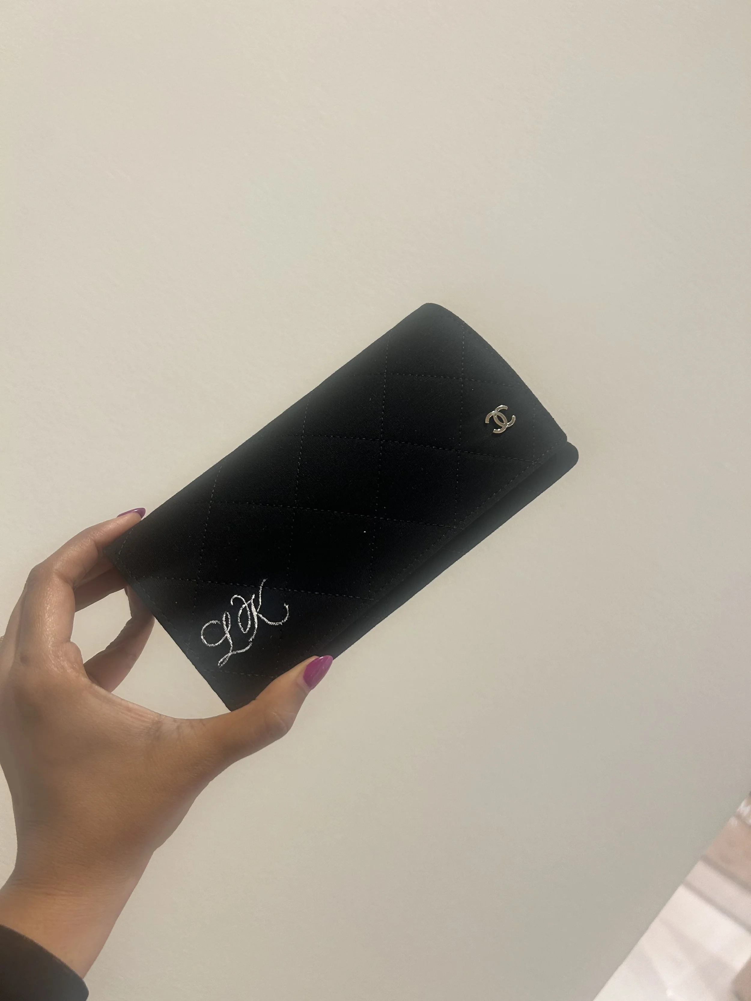 A black quilted Chanel wallet with a silver logo in the top right corner, held by a person's hand against a plain light-colored background.
