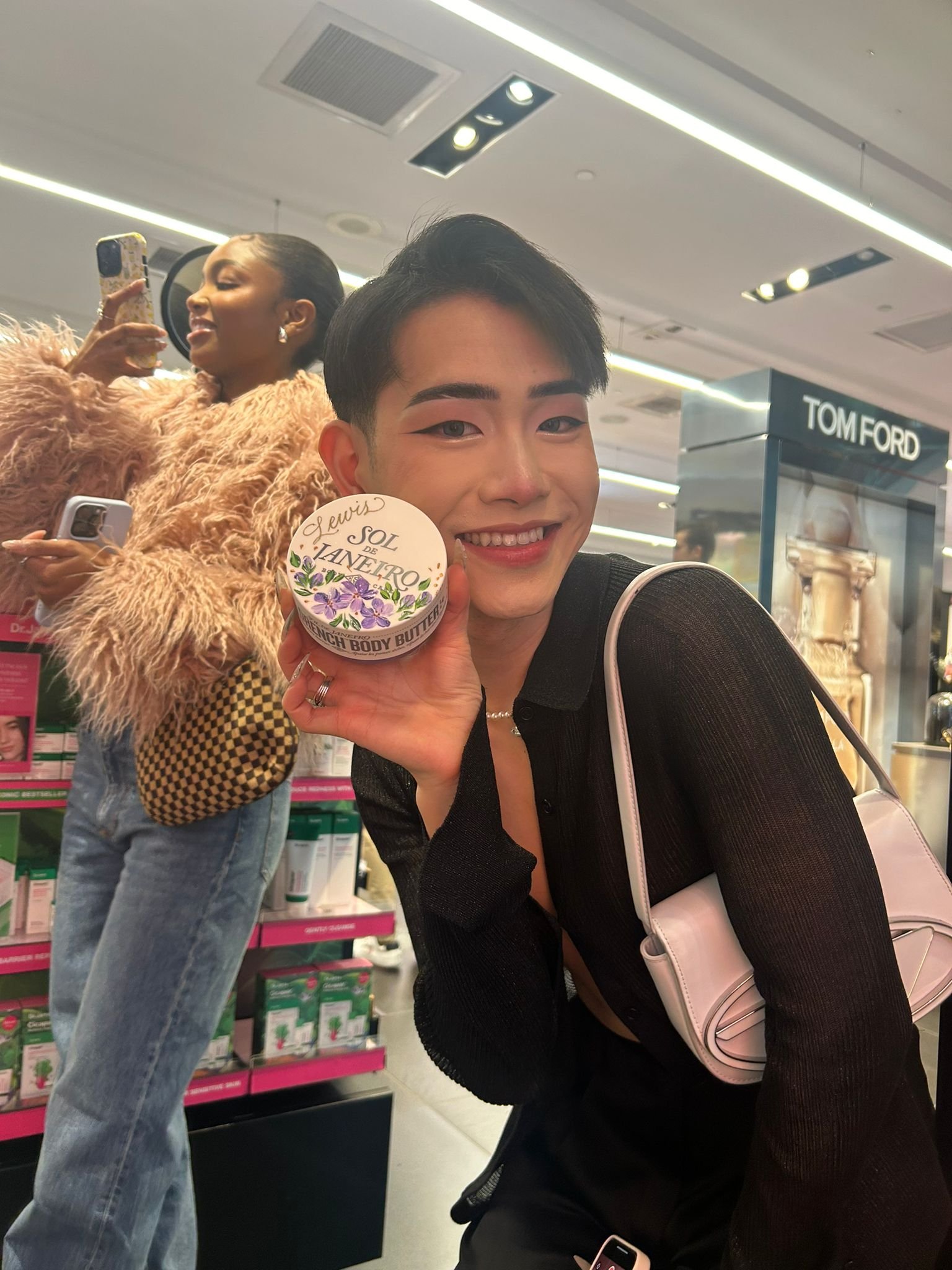 A young person smiling and holding a container of Sol de Janeiro body butter in a store. Behind them, a woman with a fluffy pink jacket and checkered bag is taking a photo with her smartphone.