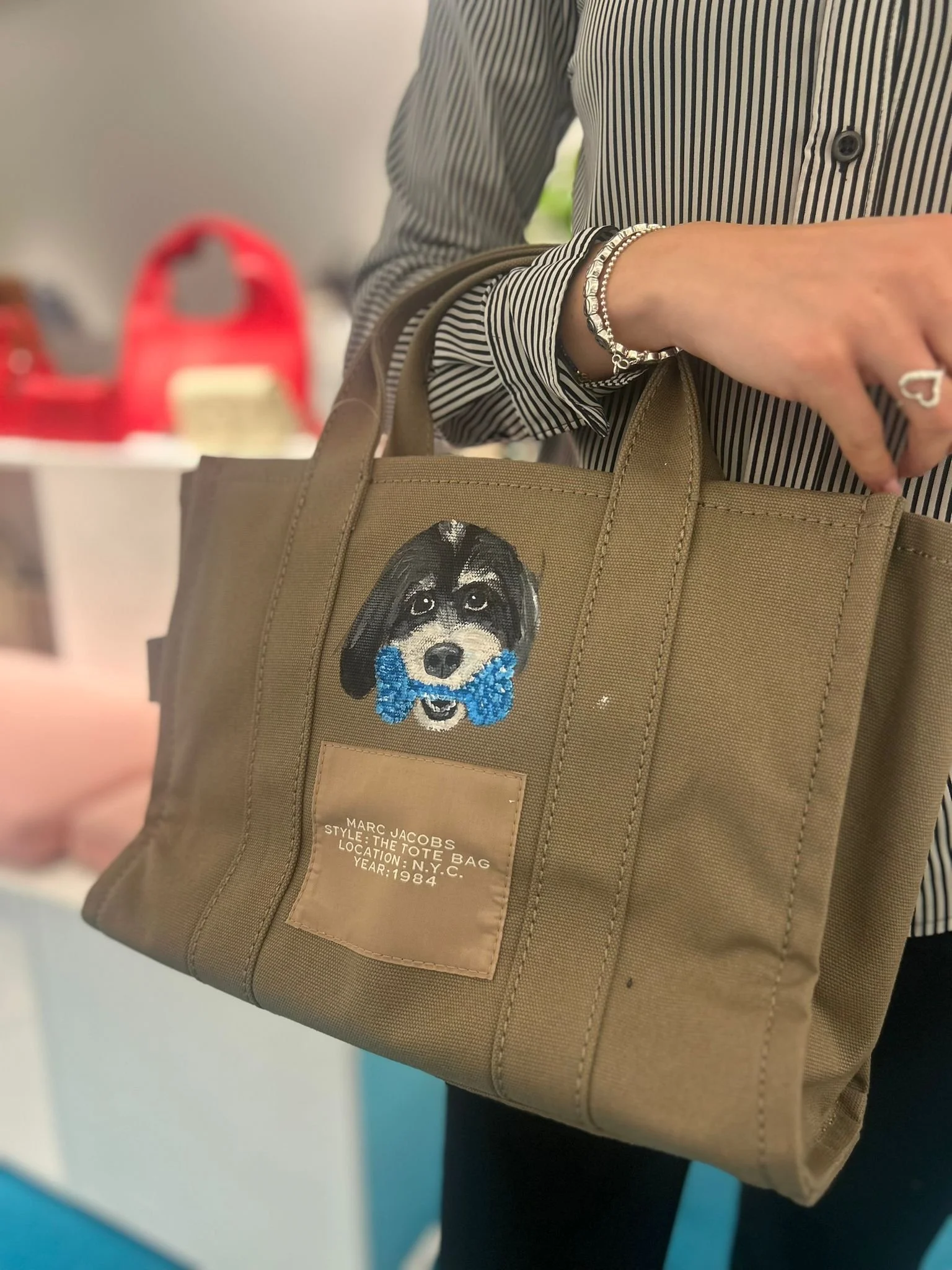 A person holding a beige tote bag with a patch of a dog's face wearing a blue bowtie. The person is wearing a silver bracelet and a ring, and the bag has a label indicating it is by Marc Jacobs, style: tote bag, from New York City, 1984.