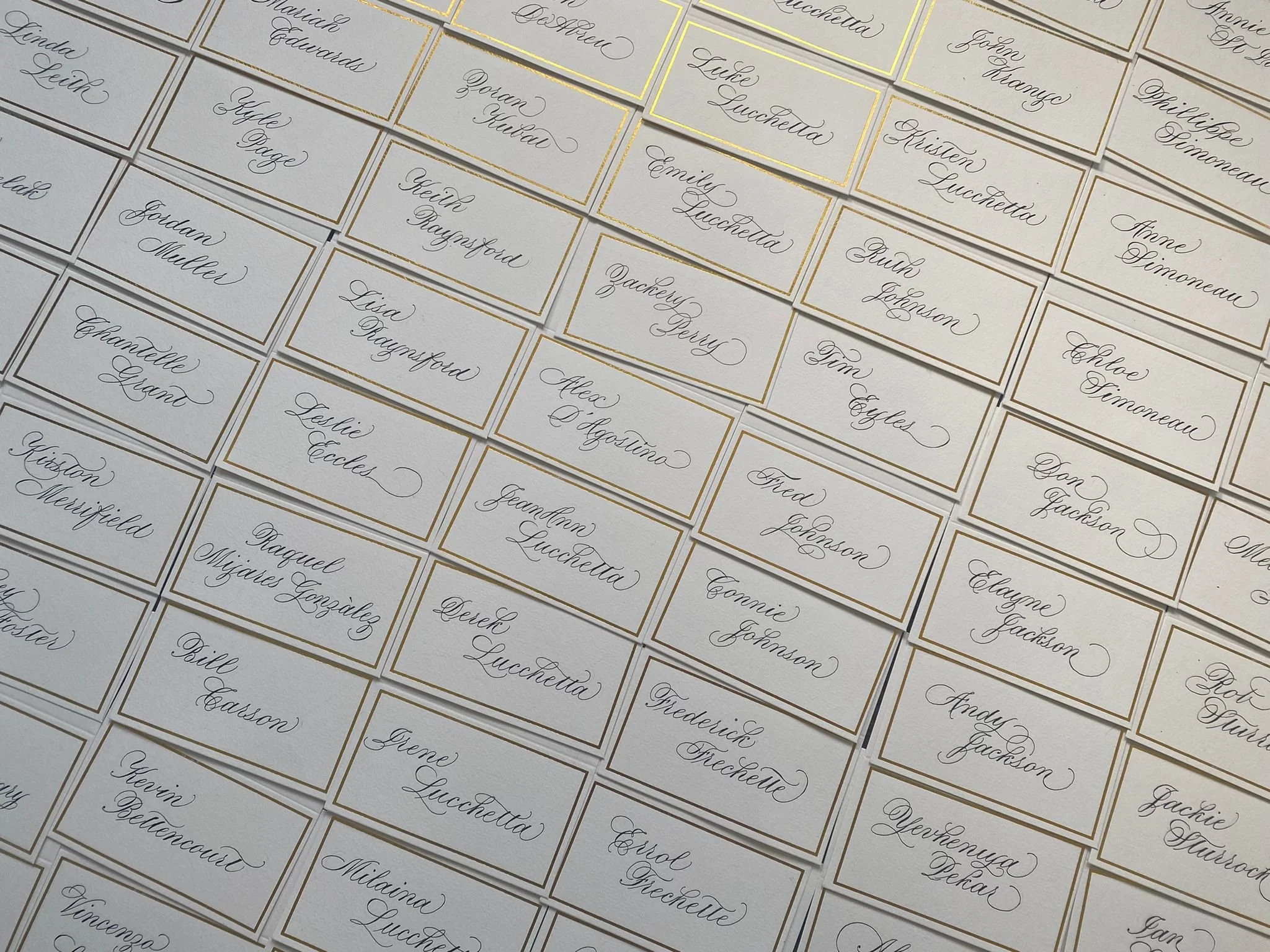 Close-up photo of multiple white place cards with handwritten names in cursive, bordered in gold, arranged in a grid pattern.