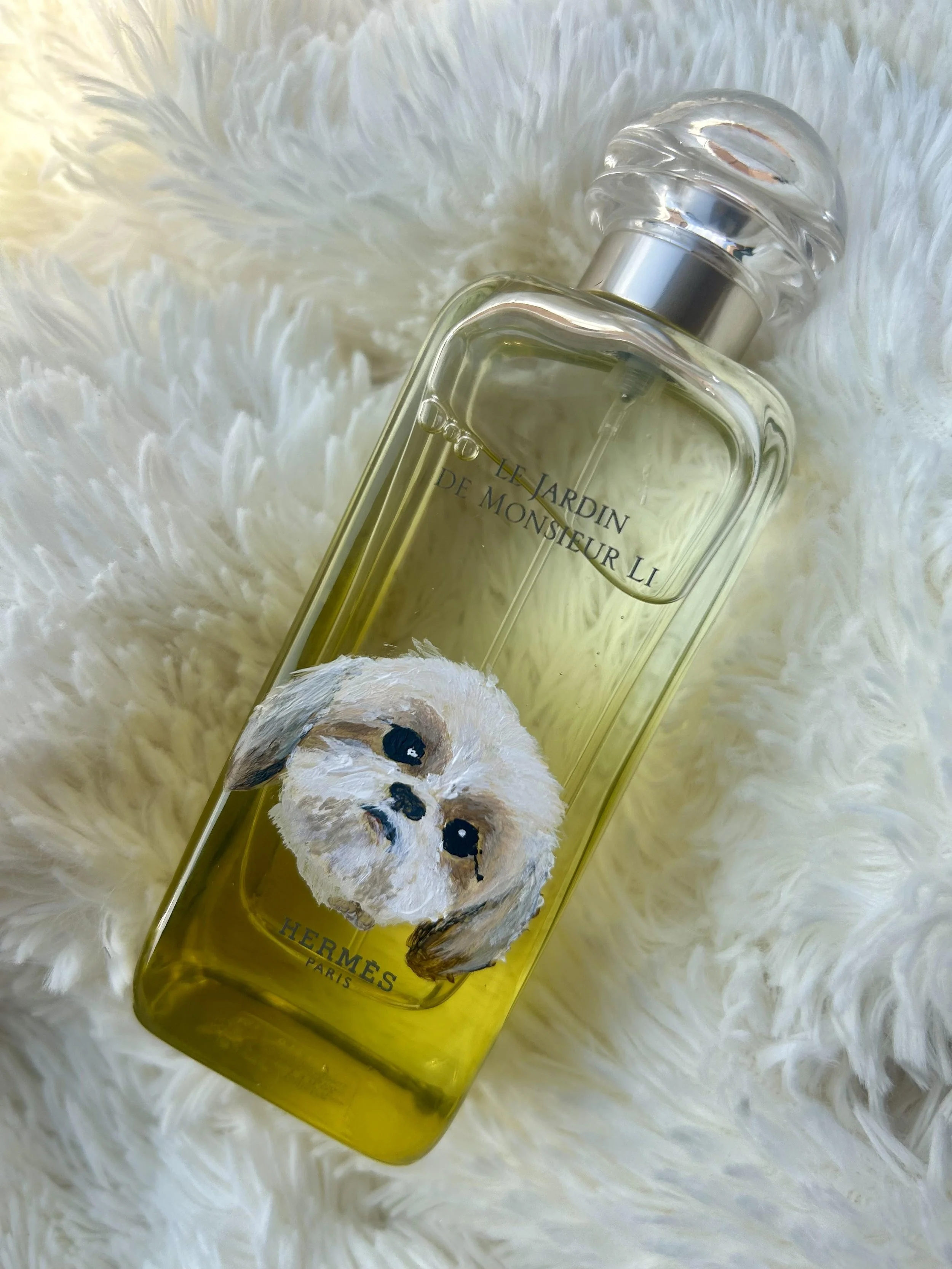 A perfume bottle labeled 'Le Jardin de Monsieur Li' by Hermès with a cute puppy illustration on a white fuzzy surface.