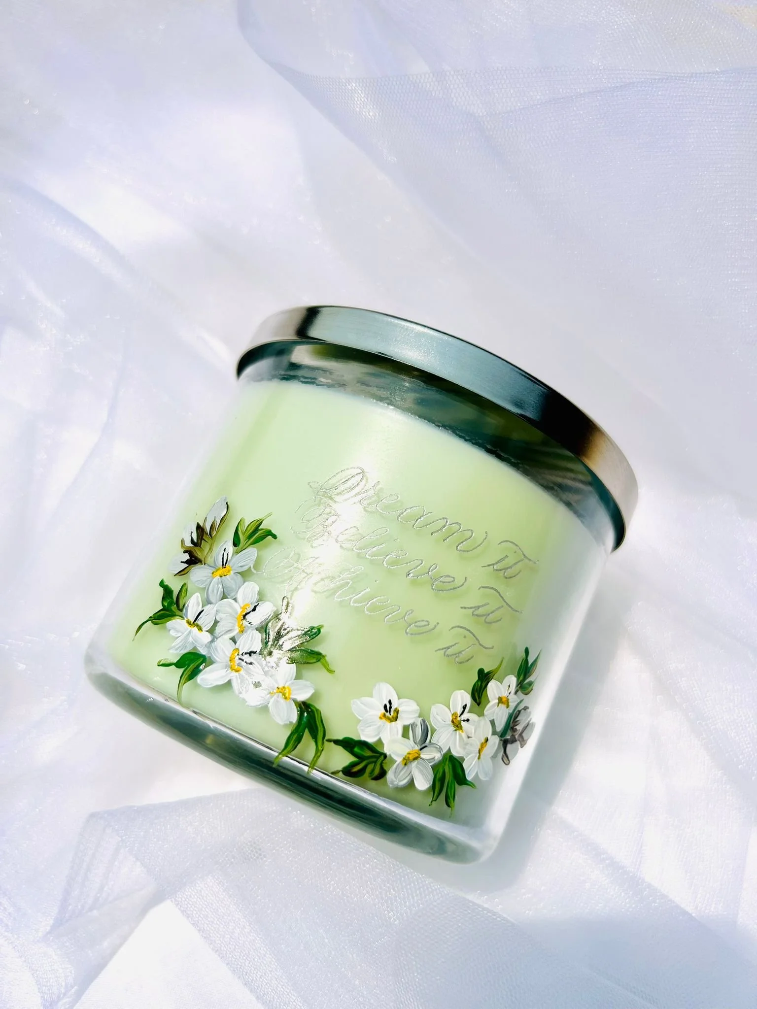 A light green candle in a glass jar with a silver lid, decorated with white flowers and green leaves, and the words "Dream It Believe It Achieve It" engraved on the front, placed on white fabric.