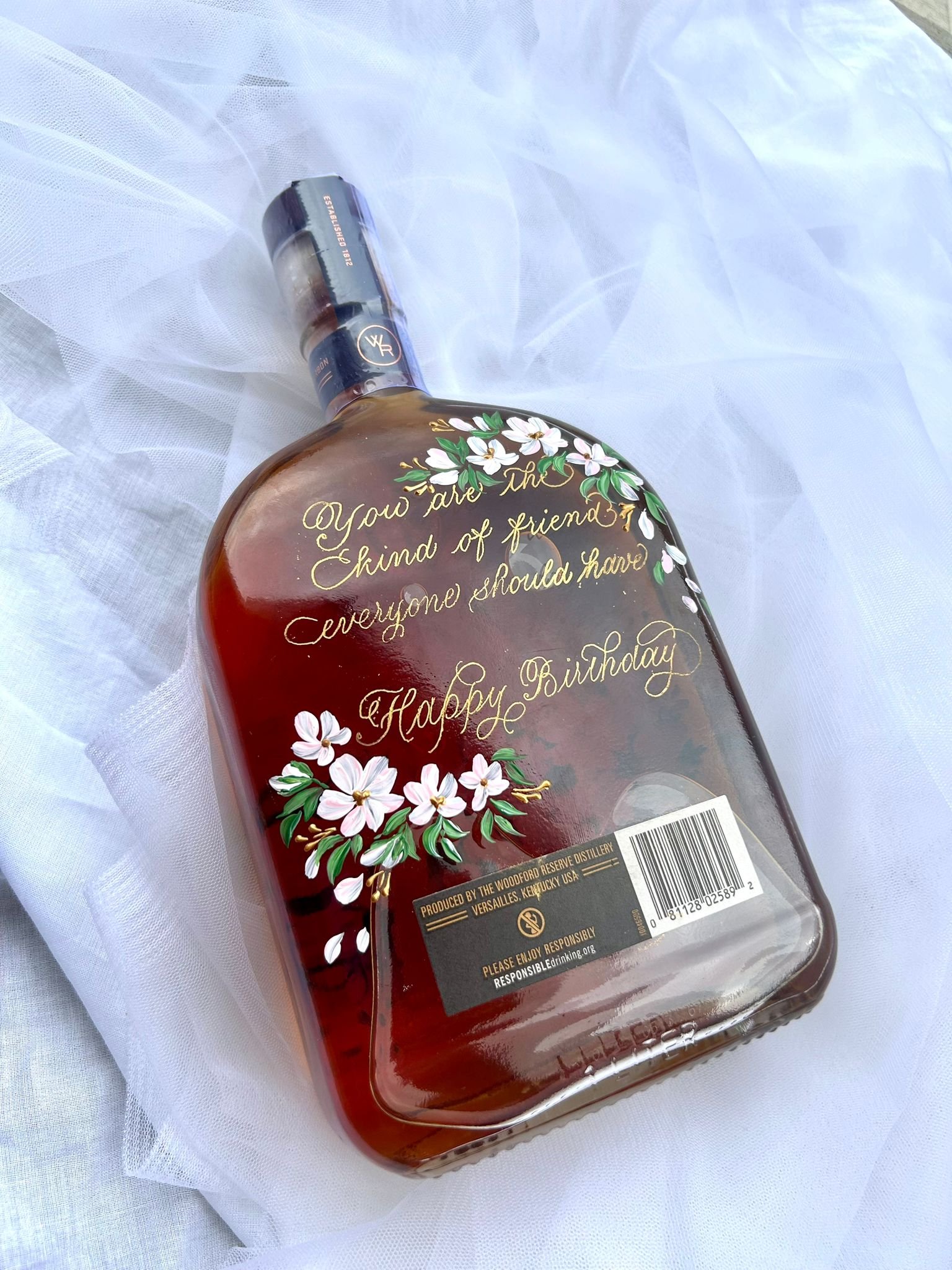 A decorated bottle with flowers and a birthday message on it, placed on white fabric.
