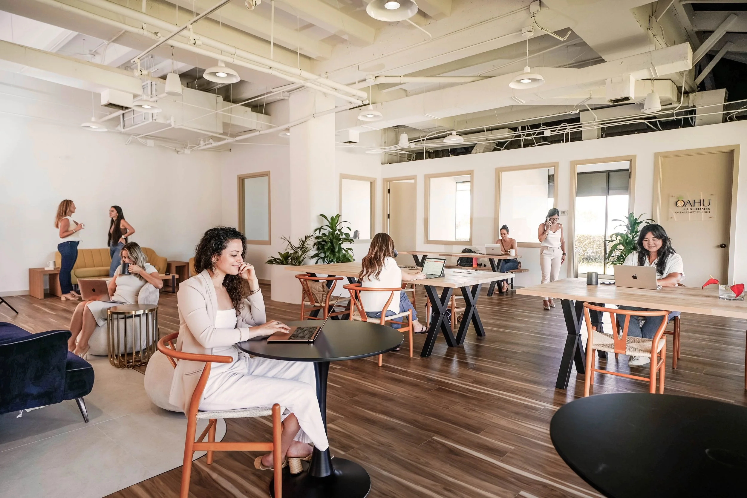 A spacious, modern office with several women working on laptops and talking on phones, some sitting at tables and others standing or walking around, with large windows and wooden floors.