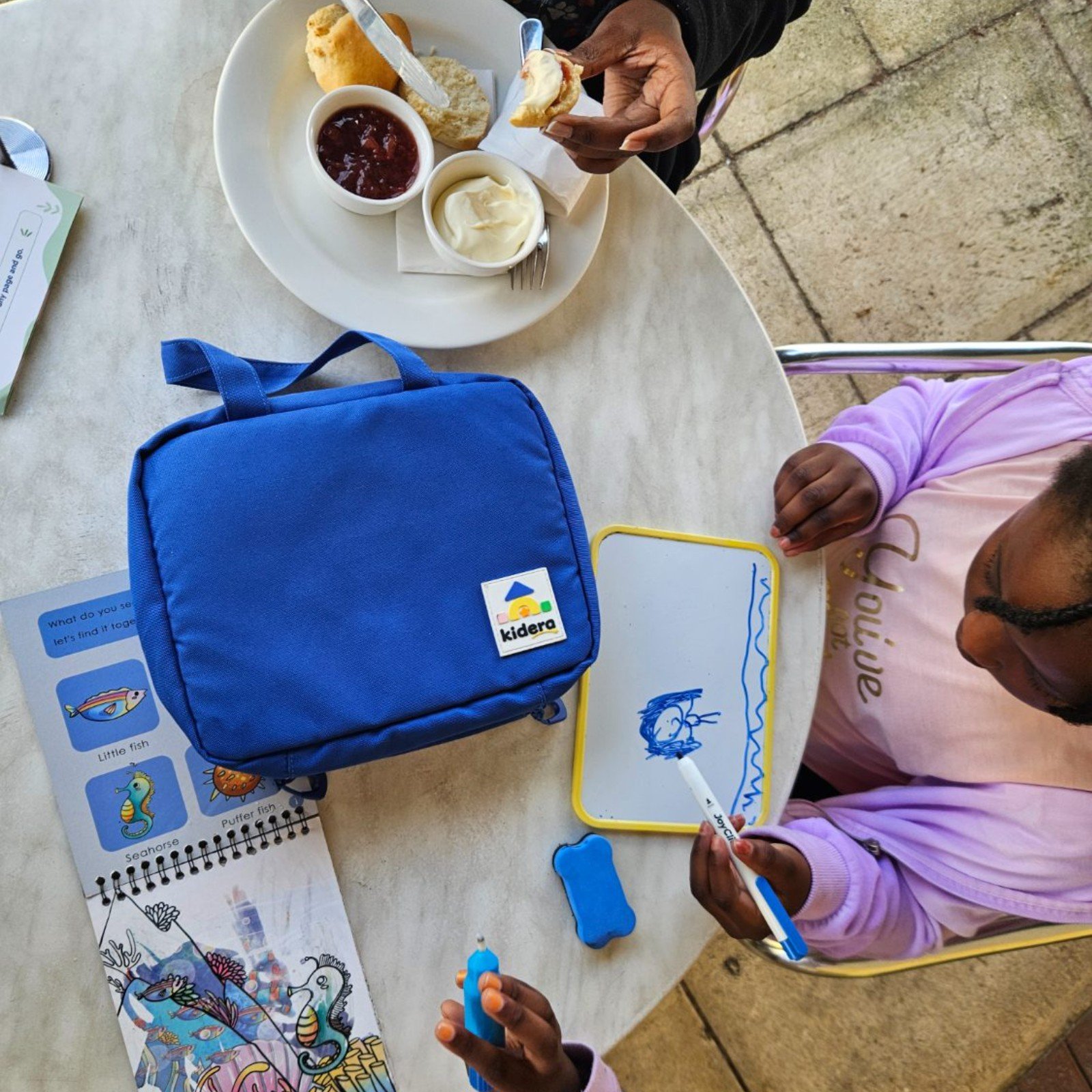 Two children drawing and colouring at a cafe table with the Kidera Travel Play Kit beside them