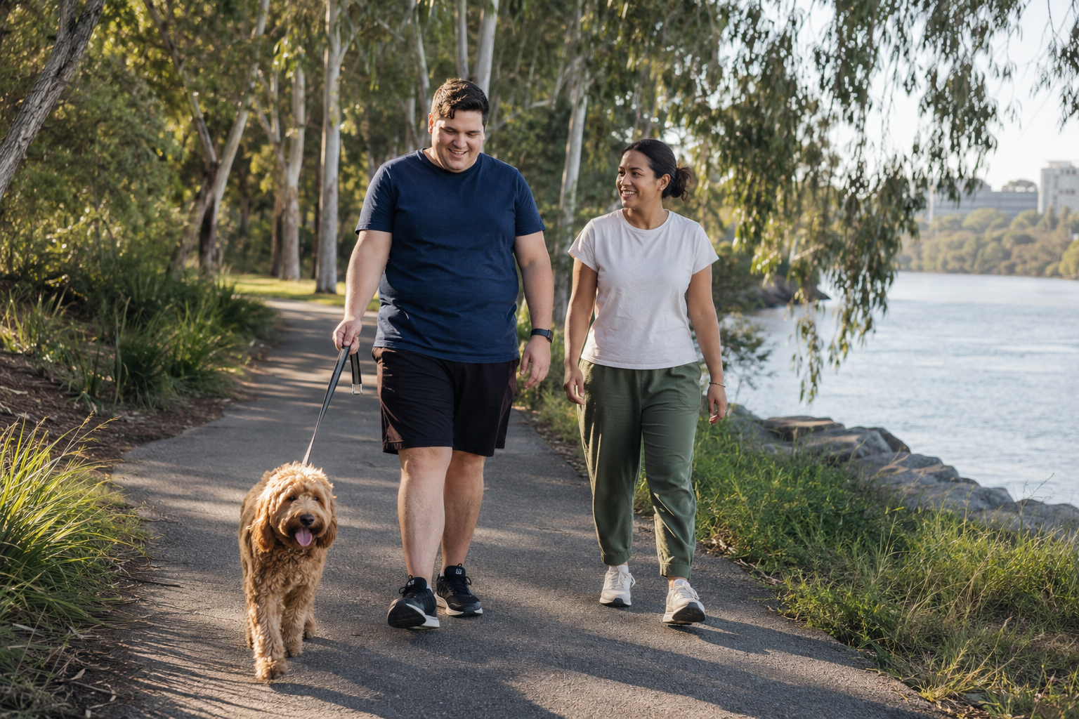 Adult participant walking a dog alongside a behaviour support practitioner on a Brisbane riverside path.
