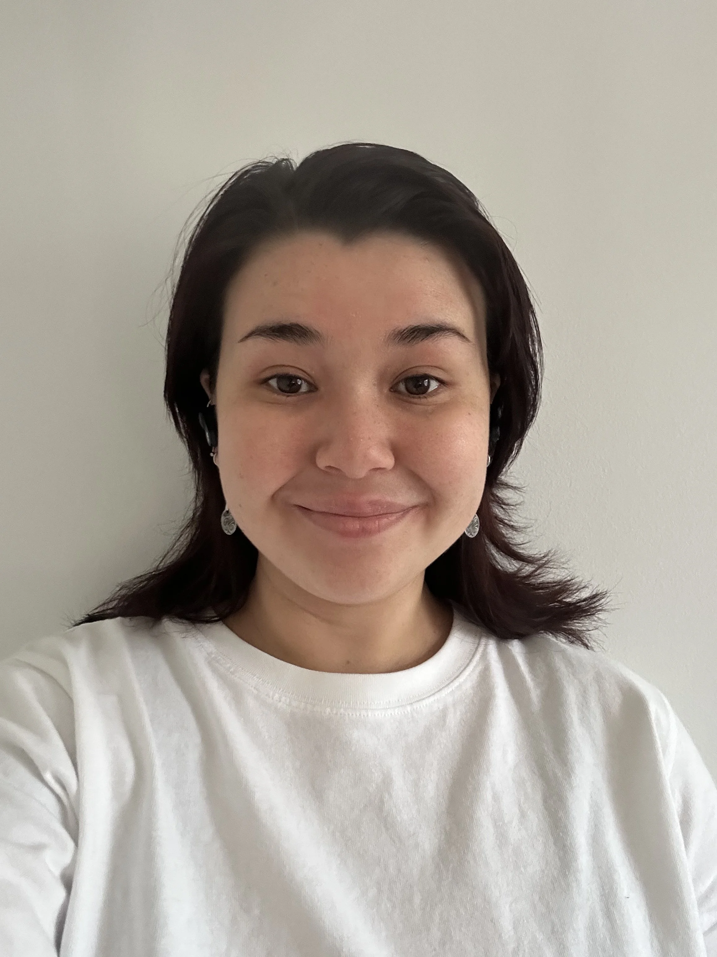 A woman with short dark hair wearing a white shirt and earrings, standing against a plain light-colored wall, smiling softly.