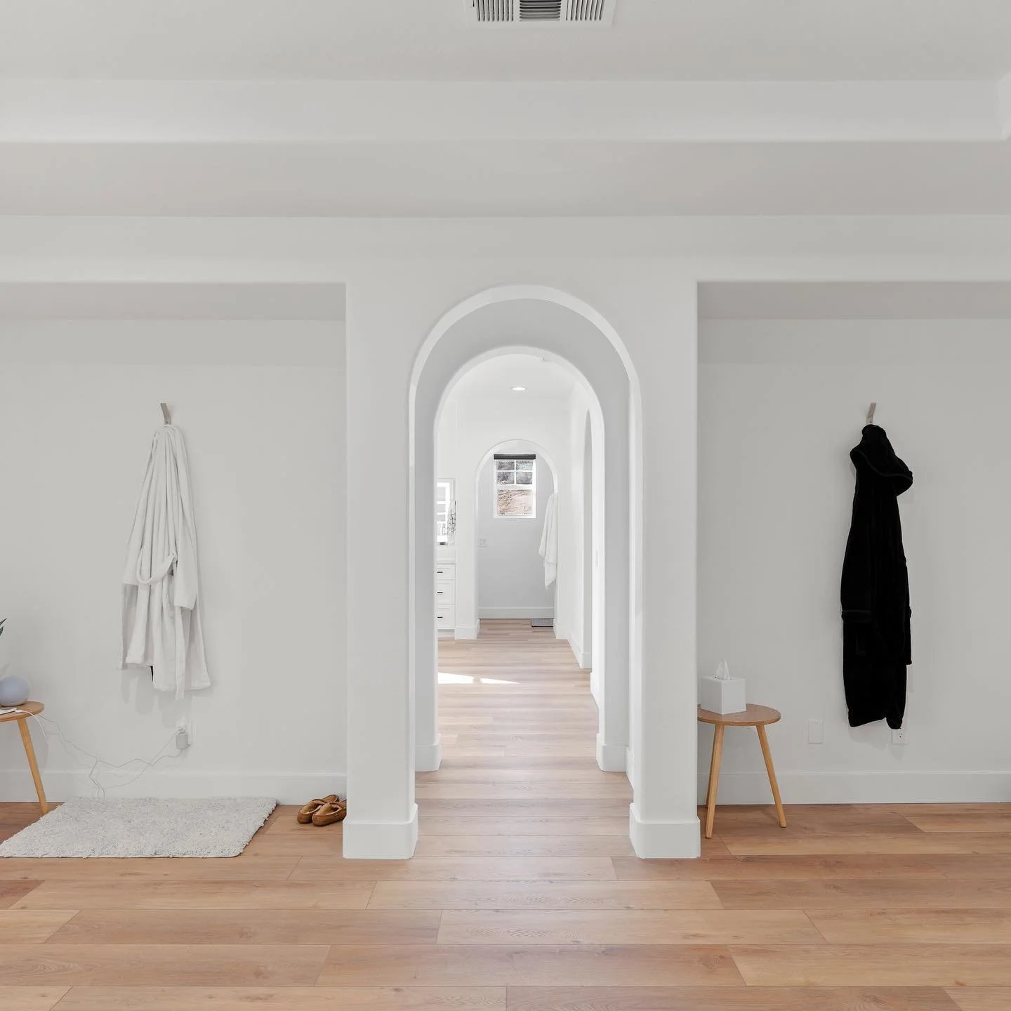 A minimalist, modern interior doorway with white walls, curved arches, and light wood flooring. On the left, a small wooden table with a potted plant, and a pair of brown slippers on a rug underneath. On the right, a black bathrobe hanging on a hook, with a small wooden side table and tissue box below. Light streams through a window at the end of the hallway.