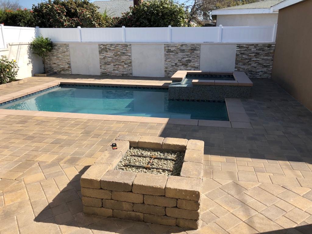 A backyard with a rectangular pool, a hot tub, a brick firepit, paver patio, and a white privacy fence with stone accents.