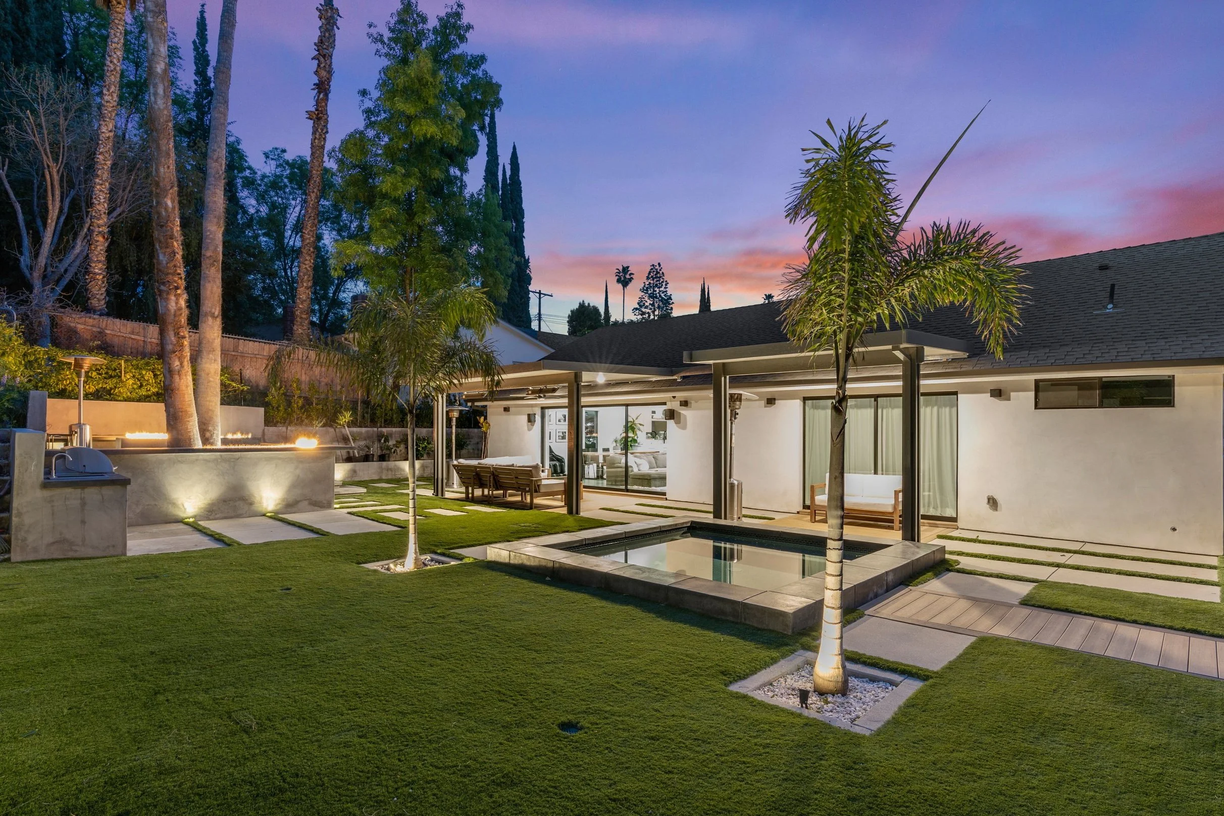 Modern backyard with a small pool, outdoor seating, palm trees, and a well-maintained lawn during sunset.