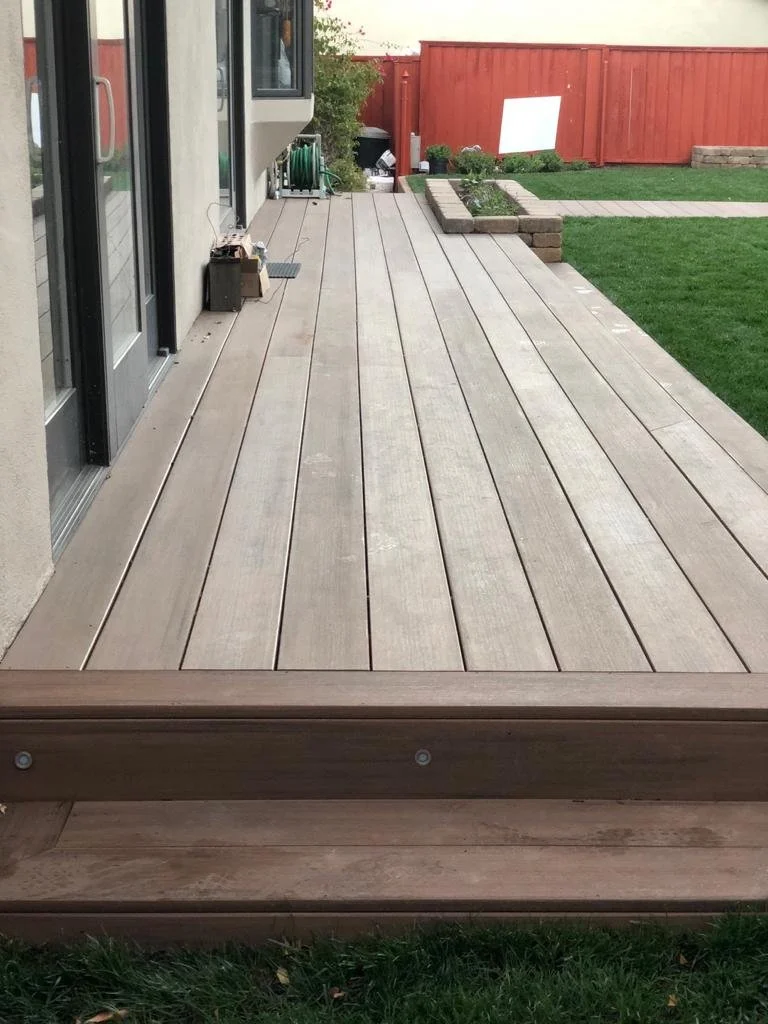 View of a wooden deck attached to a house with sliding glass doors, leading to a backyard with green grass and a red fence, raised steps at the edge of the deck, a garden area with plants and stacked bricks, and outdoor storage and equipment.