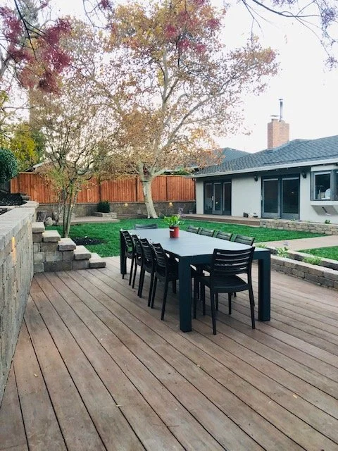 Backyard patio area with a wooden deck, a black dining table with six chairs, a red potted plant, green grass, and a house with a chimney in the background.