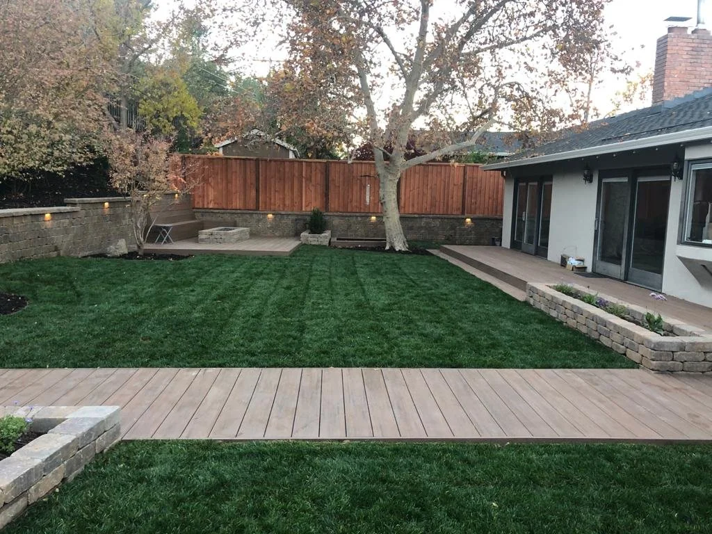 Backyard with a lush green lawn, a large central tree, a wooden deck, a paved walkway, and a house with large glass doors and windows.
