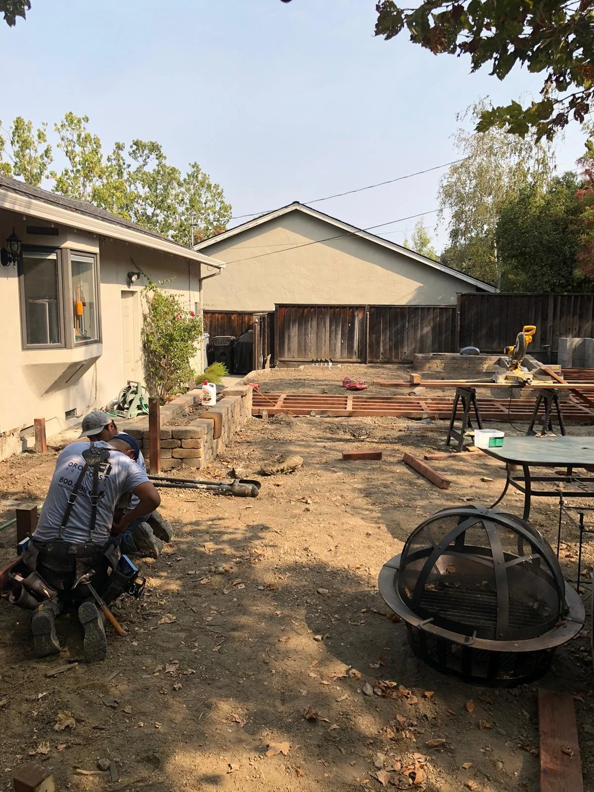 Construction workers working on a backyard renovation. The area is partially excavated with construction tools and wooden framing for a deck or patio. A fire pit is visible in the foreground, and there is a house with a window on the left side of the image. A wooden fence surrounds the backyard.