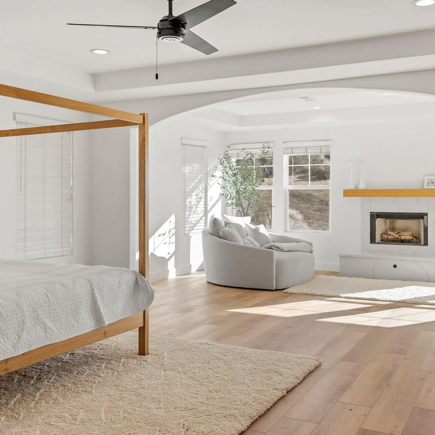 Bright living room with white walls, wooden floor, a bed frame on the left, a white sofa by the window, fireplace with wooden mantel, and a potted plant, all illuminated by natural light.