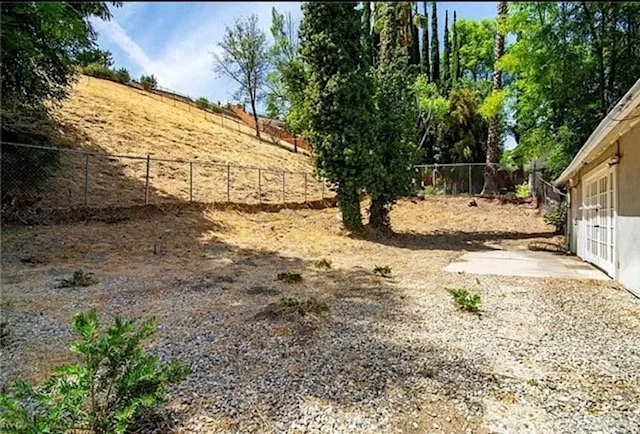A backyard yard with a gravel ground, a large tree, and a house with a garage on the right. There is a wooden fence along the back and a sloped, grassy area with a brick wall on the left.