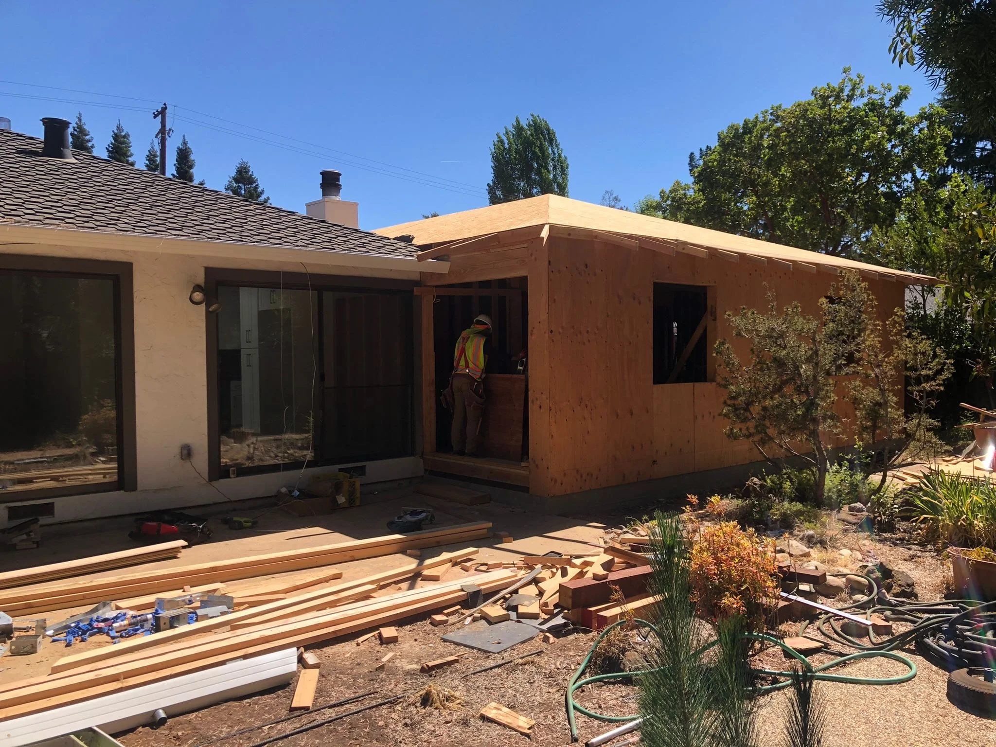 Construction worker building a wooden extension on a house under a blue sky, with tools and building materials scattered on the ground.