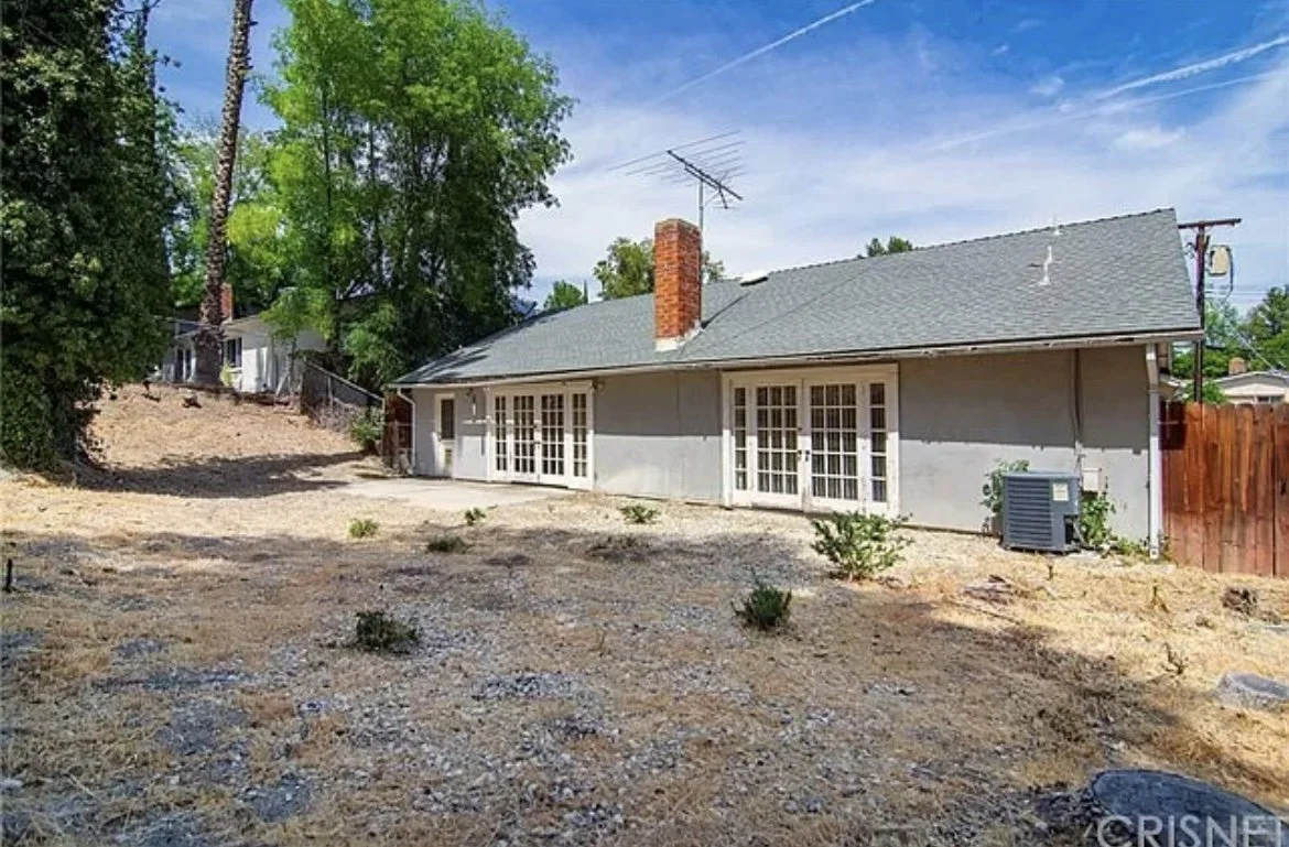 Backyard of a house with a gravel dirt ground, a few small plants, and a gray house with a brick chimney and French doors, surrounded by trees and neighboring houses.
